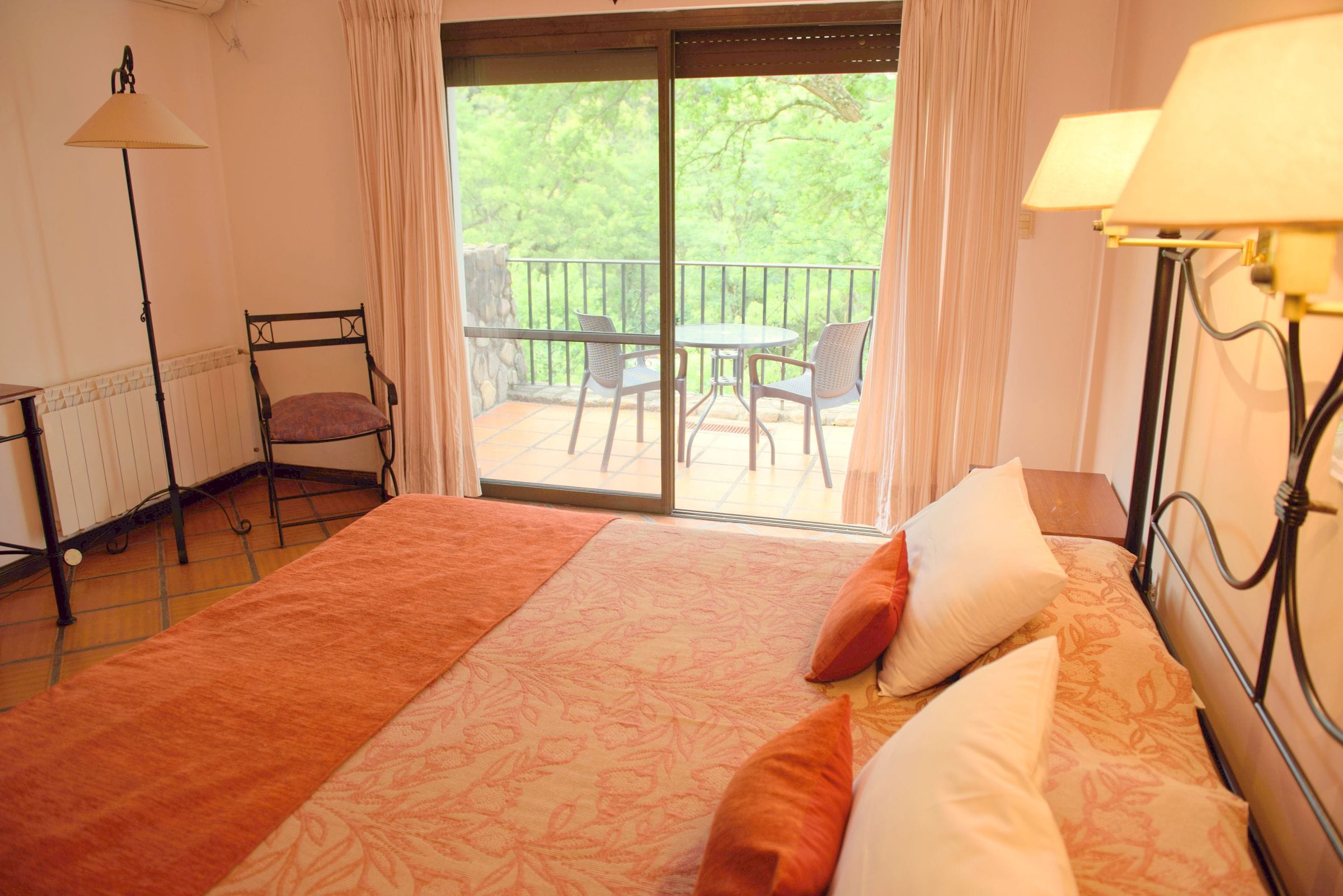 A bedroom with a large bed, a balcony with chairs, and a view of greenery.