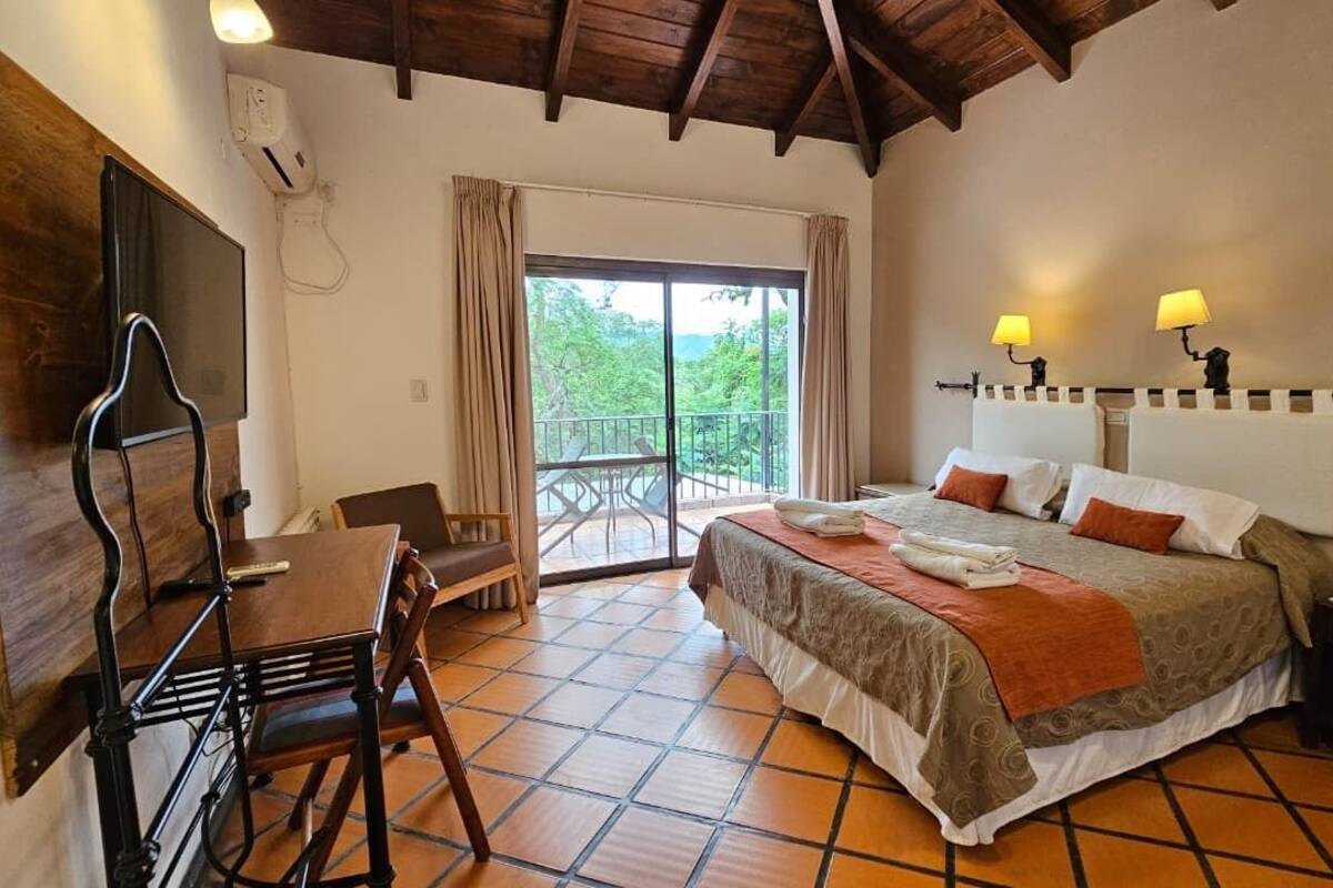A bedroom with a bed, a desk, and a chair. There is a balcony with a table and chairs.