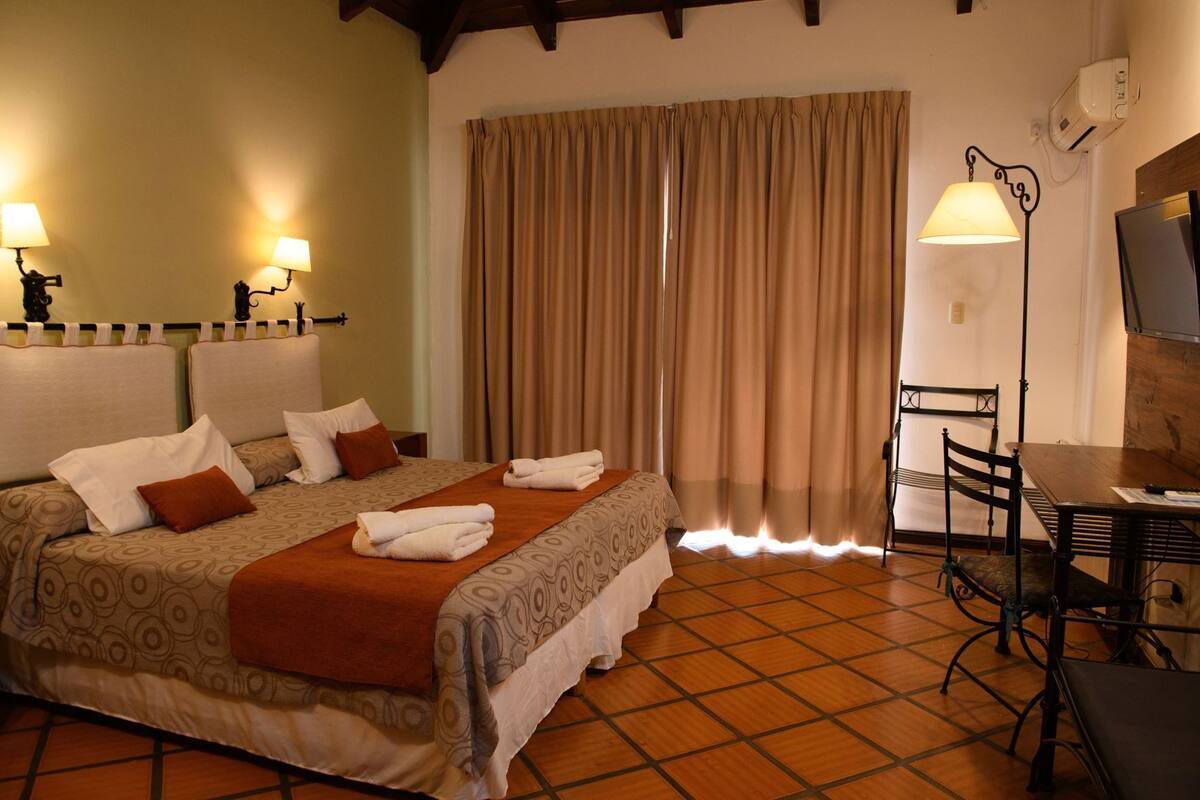 A hotel room with a bed, two bedside lamps, a desk with a chair, a television, and a window with curtains.