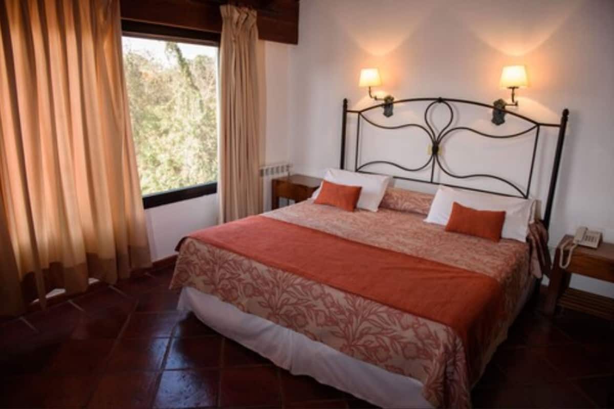 A neatly made bed with a patterned bedspread and pillows, a nightstand with a lamp, and a window with curtains.
