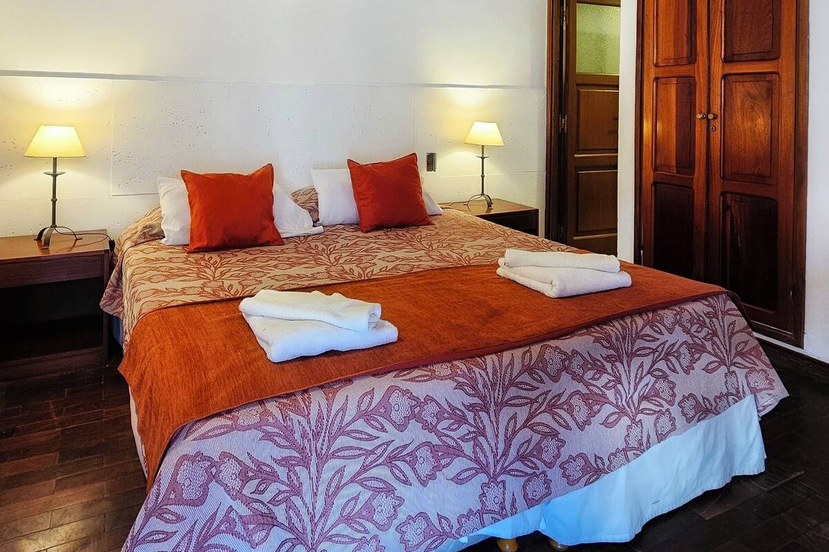 A bedroom with a wooden ceiling, a bed with a patterned bedspread, two bedside lamps, and a wooden wardrobe.