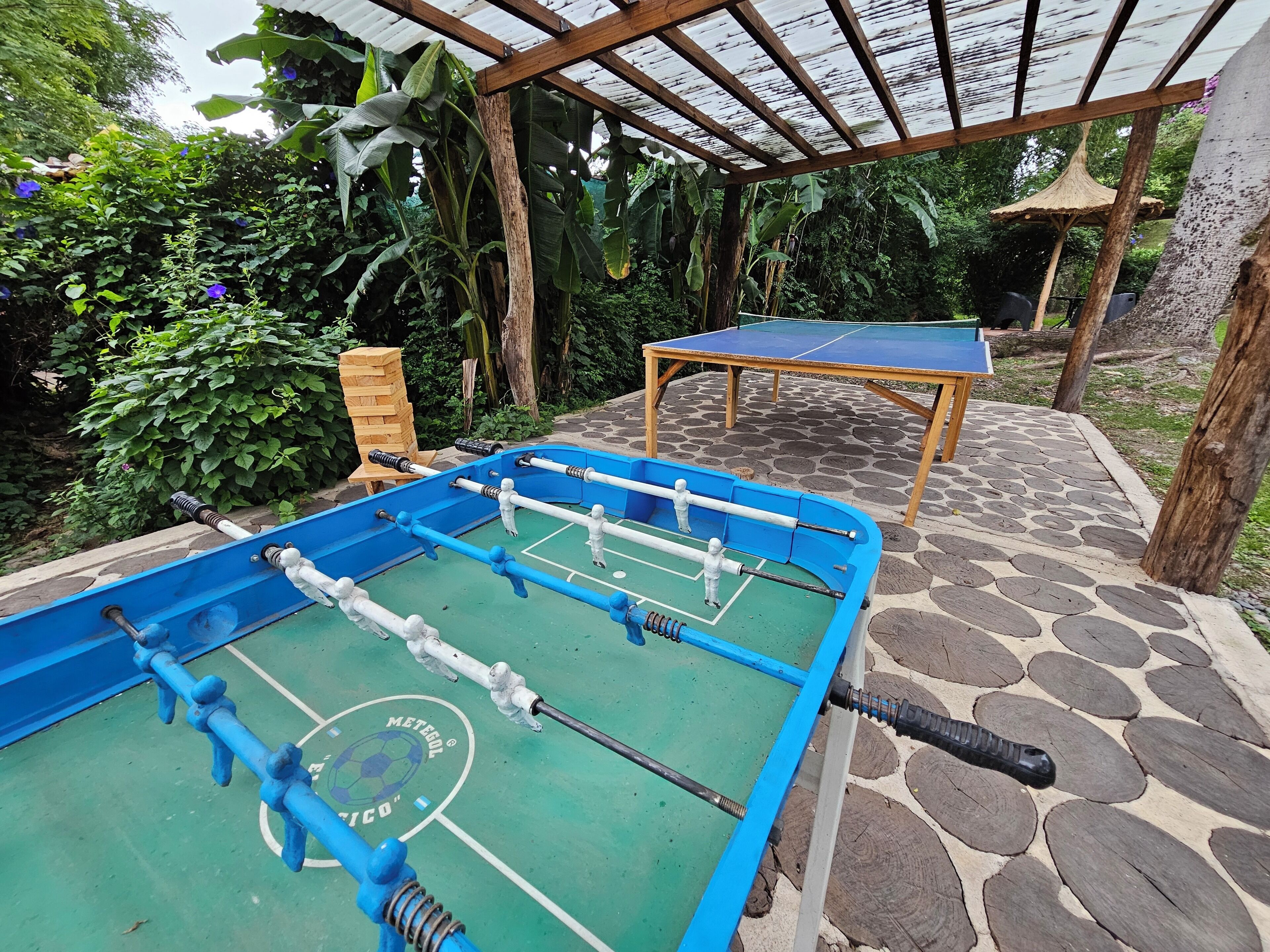 Taula de futbolí i taula de ping-pong en una zona exterior amb una pèrgola de fusta a la part superior.