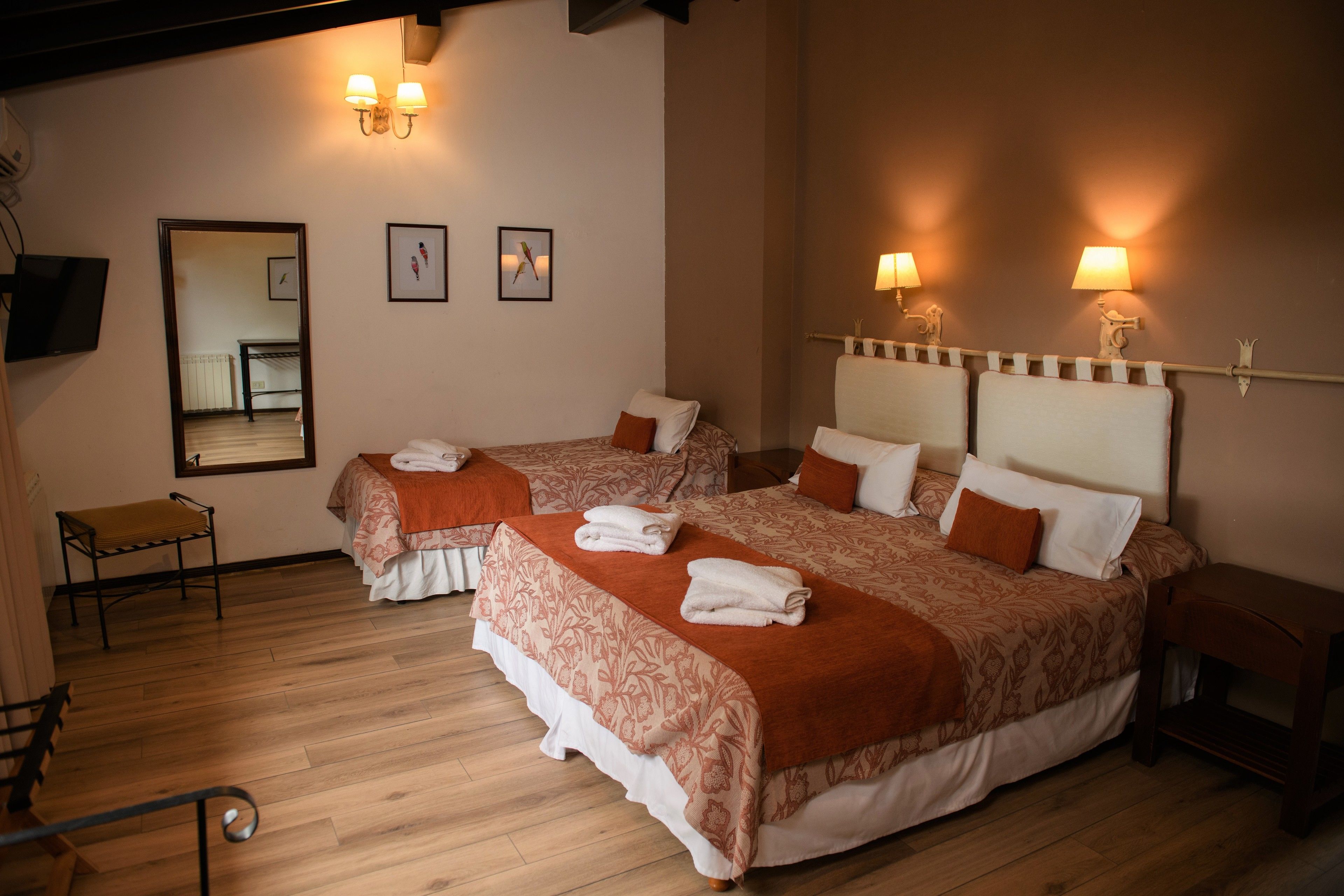 A hotel room with two beds, a small table, and a bench. There are wall-mounted lamps and framed pictures.
