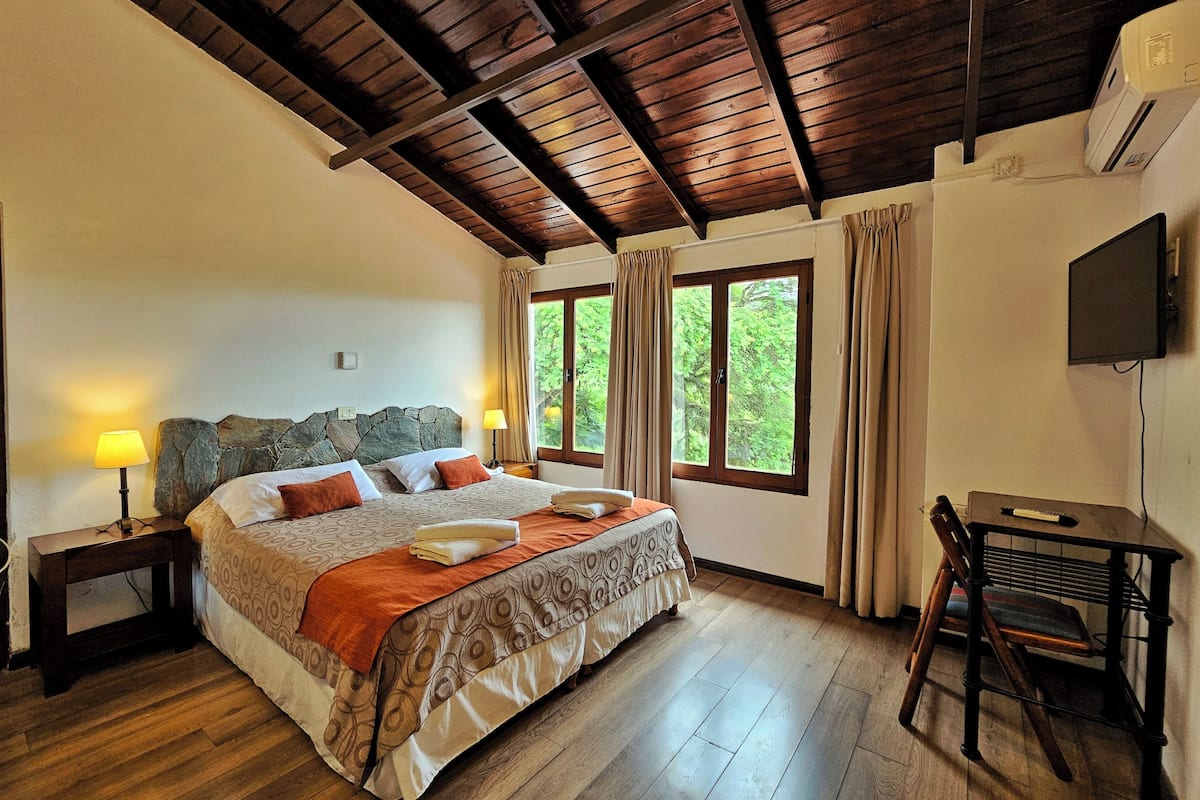A bedroom with a large bed, wooden flooring, a stone headboard, a desk with a chair, and a TV on the wall.