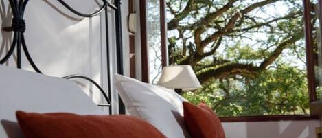 Un dormitorio con cama, almohadas, una mesita de noche, lámpara y ventana con vista a árboles.