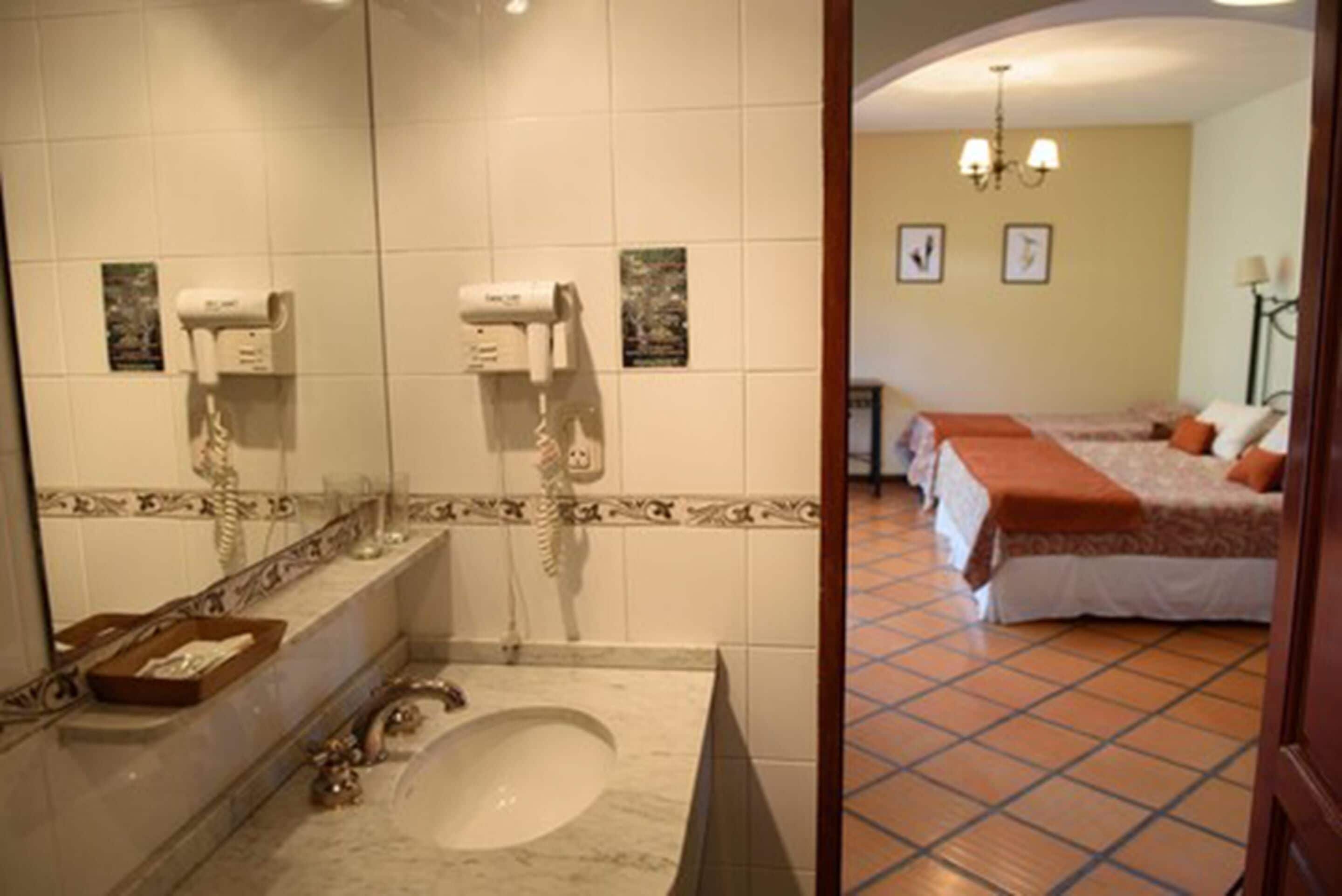 A bathroom with a marble sink, two hair dryers, and a mirror. The room has a tiled floor and a bed with a red blanket.