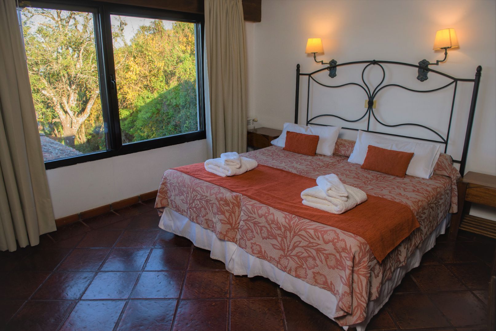 Una cama bien tendida con una colcha estampada con flores, dos lámparas de noche y vistas a árboles a través de una ventana.