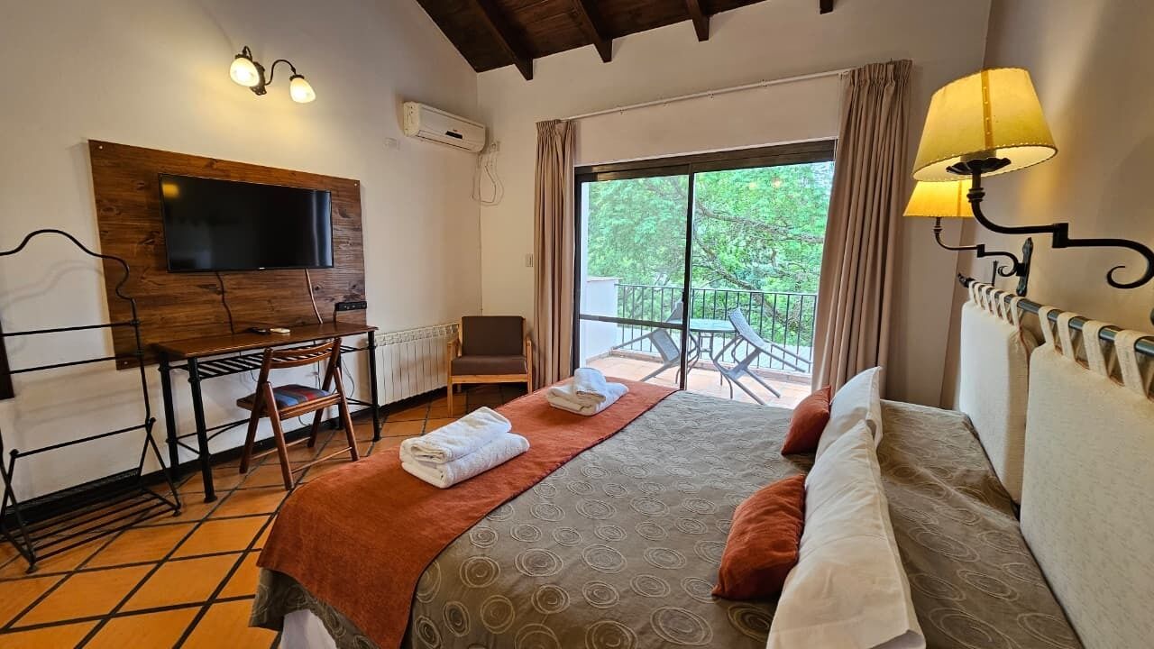 A hotel room with a bed, a desk, a chair, a TV mounted on the wall, and a balcony with a view of greenery.