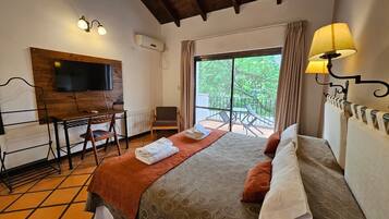 A hotel room with a bed, a desk, a chair, a TV mounted on the wall, and a balcony with a view of greenery.