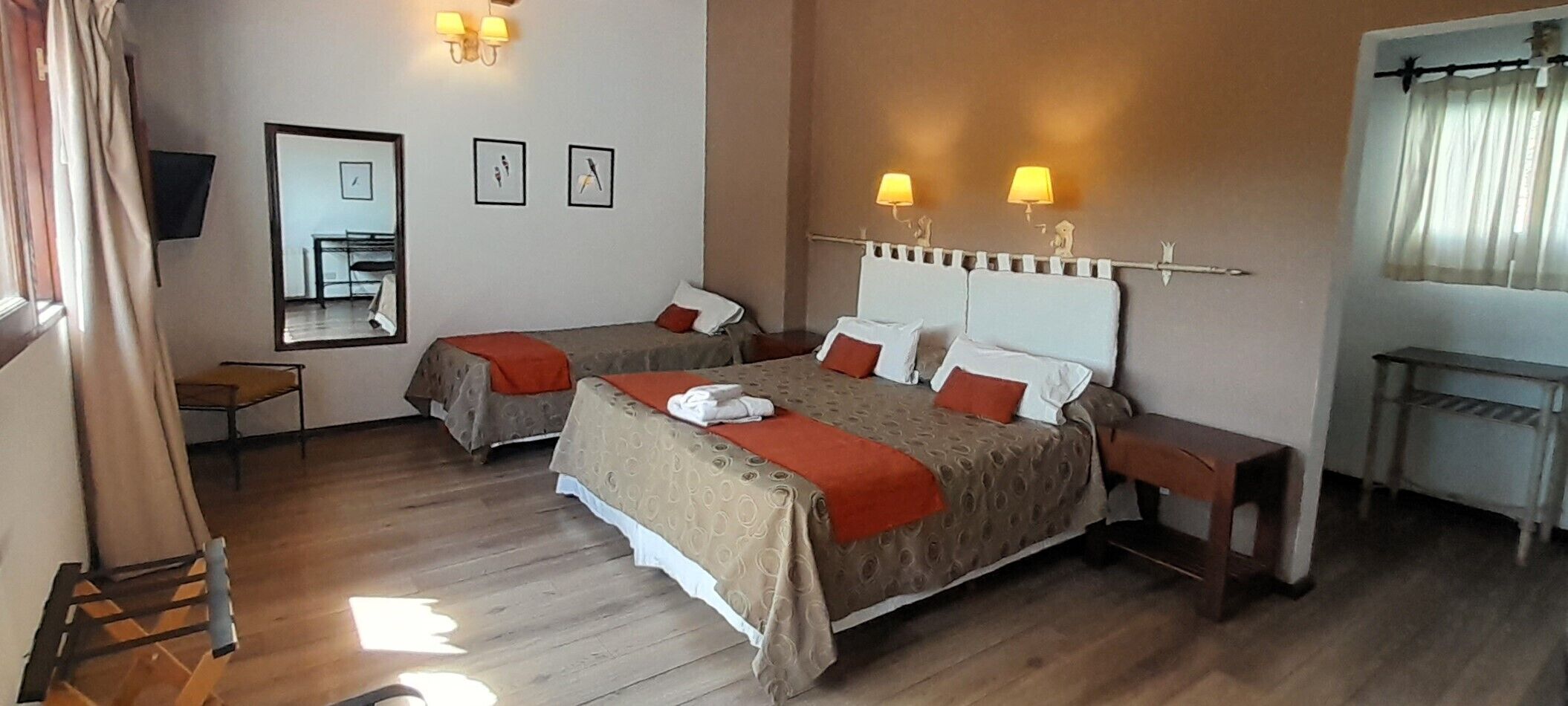A hotel room with two beds, a small table, a mirror, and a window with curtains.