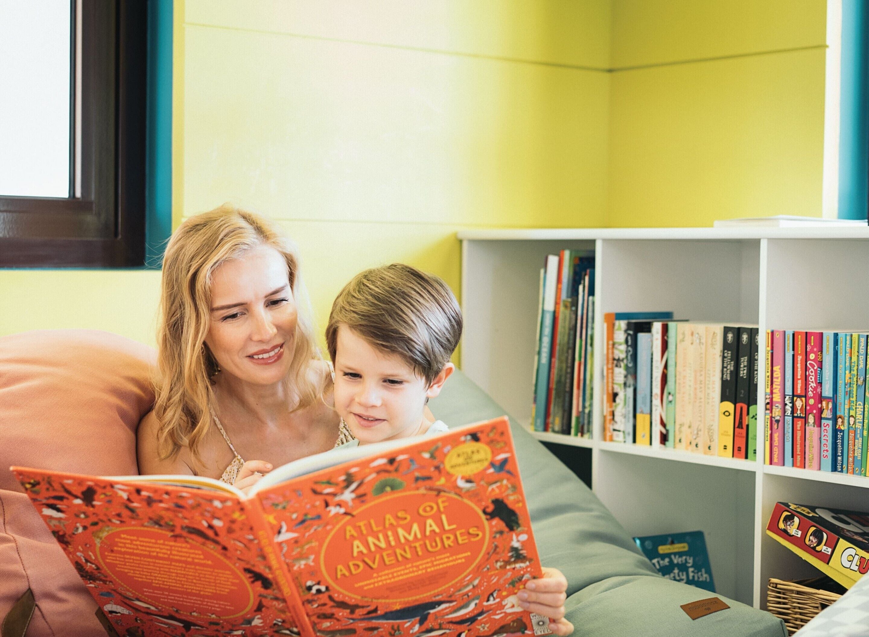 Een kamer met een boekenplank vol boeken en een kind dat een boek leest getiteld 'Atlas van dierlijke avonturen'.