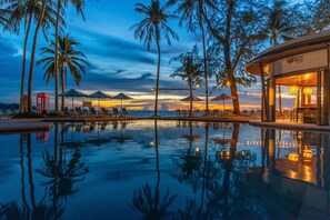 Un área junto a la piscina con sillones de descanso y sombrillas, palmeras y un edificio de diseño moderno.
