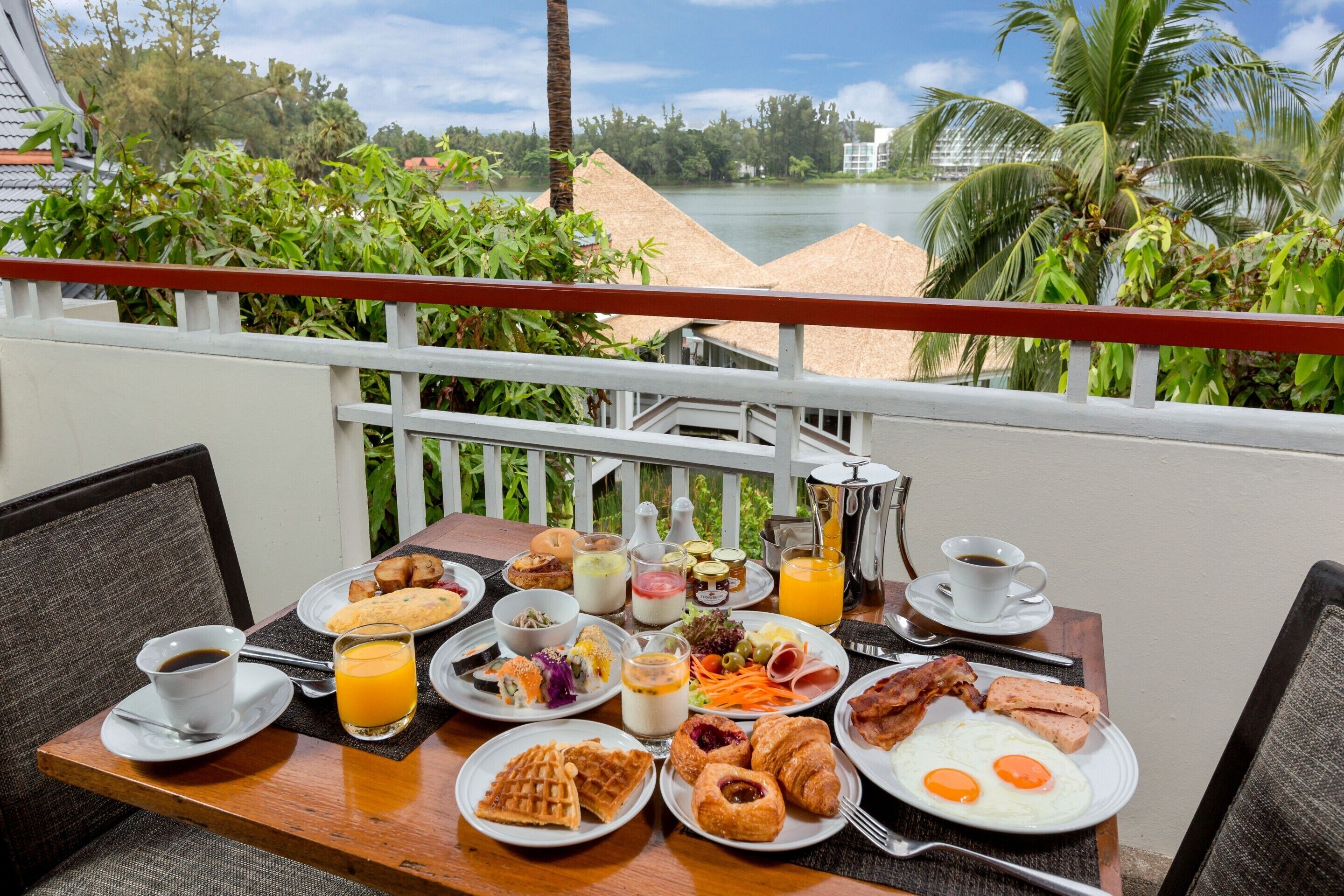 A breakfast table with various dishes and drinks, overlooking a scenic view with palm trees and a body of water.