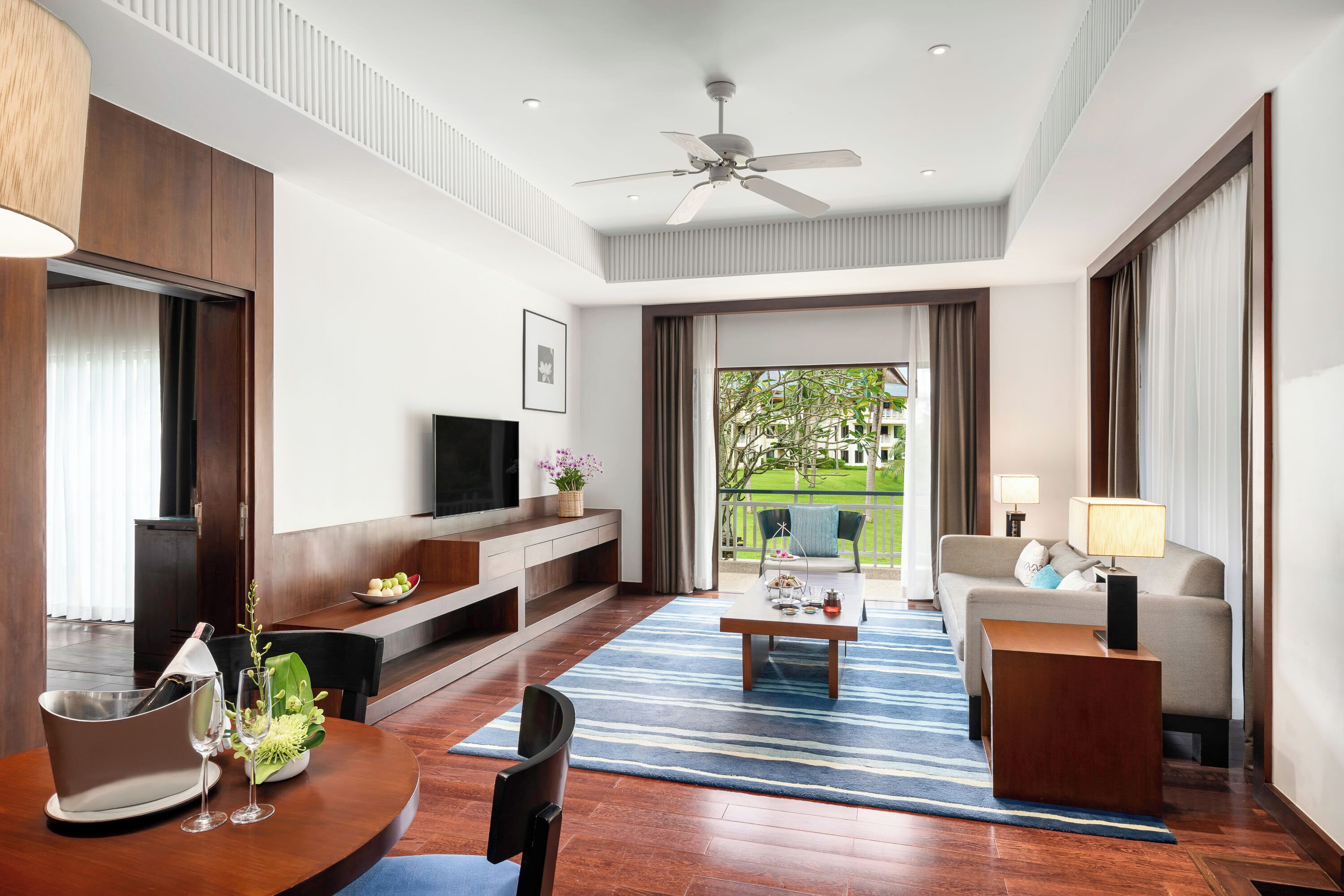A modern hotel room with a flat-screen TV, a sofa, a dining table with a blue striped rug, and a balcony with a view of greenery.