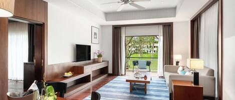 A modern hotel room with a flat-screen TV, a sofa, a dining table with a blue striped rug, and a balcony with a view of greenery.