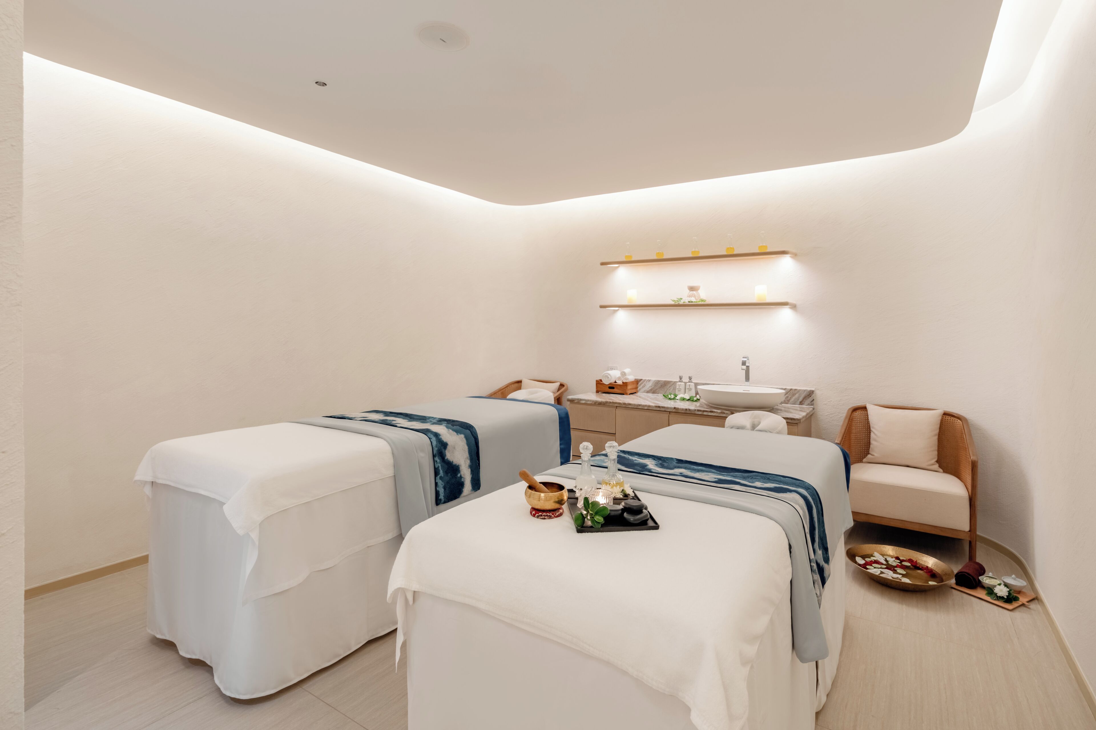 A modern spa room with two massage beds, a small table with a bowl and oils, a sink, and a shelf with toiletries.