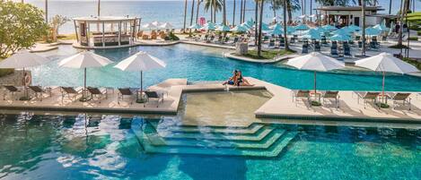 Zona de piscina en un resort con varias secciones, sillas de descanso, sombrillas y vista al océano.