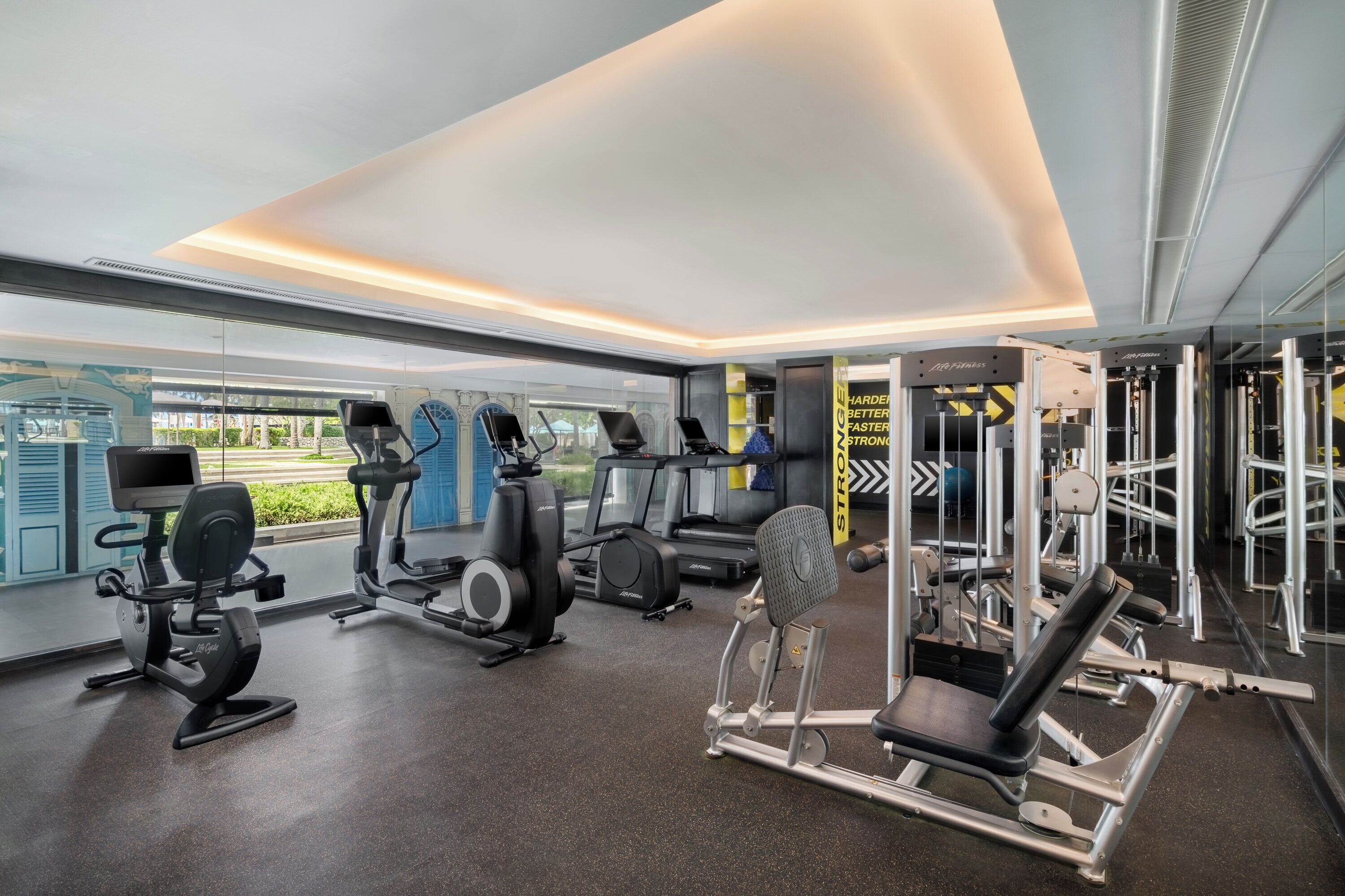 A modern gym with cardio machines, weightlifting equipment, and large windows offering a view of the outdoors.