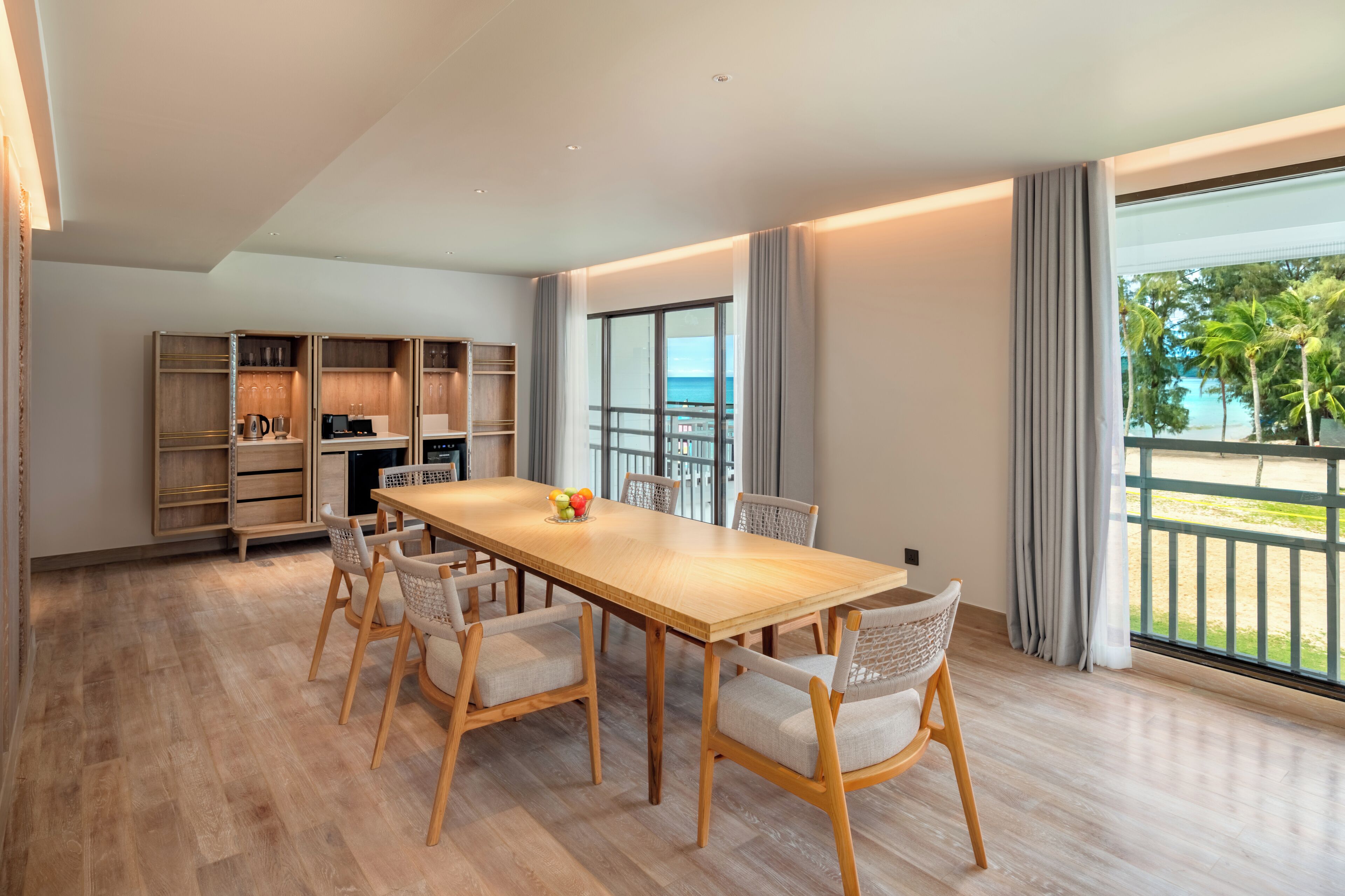 Un comedor amplio con una larga mesa de madera y seis sillas, una cocina con estantes abiertos y un balcón con vistas a palmeras.