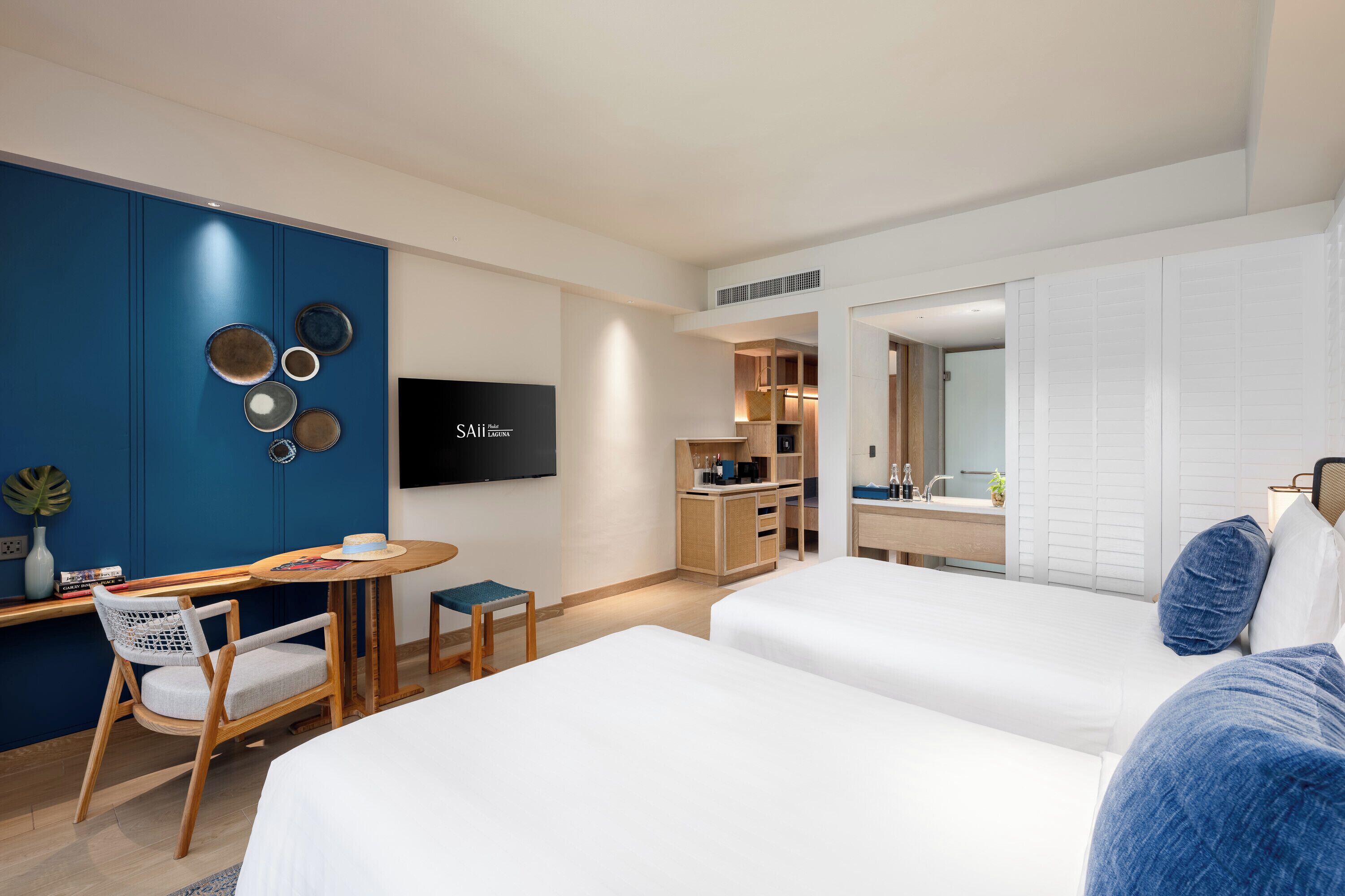 A modern hotel room with two beds, a desk with a chair, a TV, and a kitchenette.