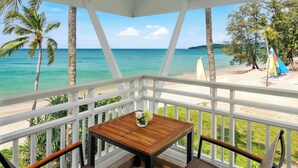 Una mesa de madera con sillas en un balcón con vista a una playa con palmeras y una vista despejada del océano.