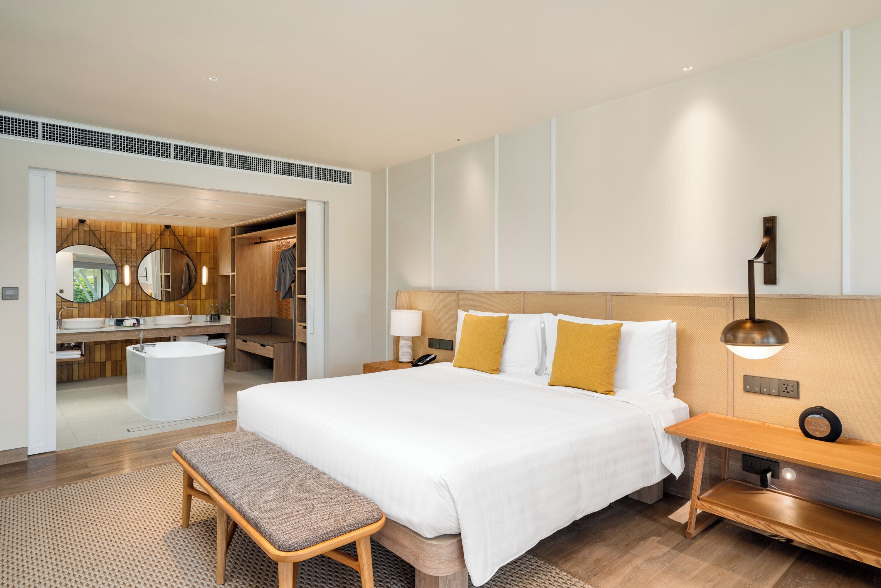 Ein modernes Hotelzimmer mit einem großen Bett, Nachttischen, einer Bank, einem Badezimmer mit Waschbecken und Spiegel sowie einer Holzwandverkleidung.