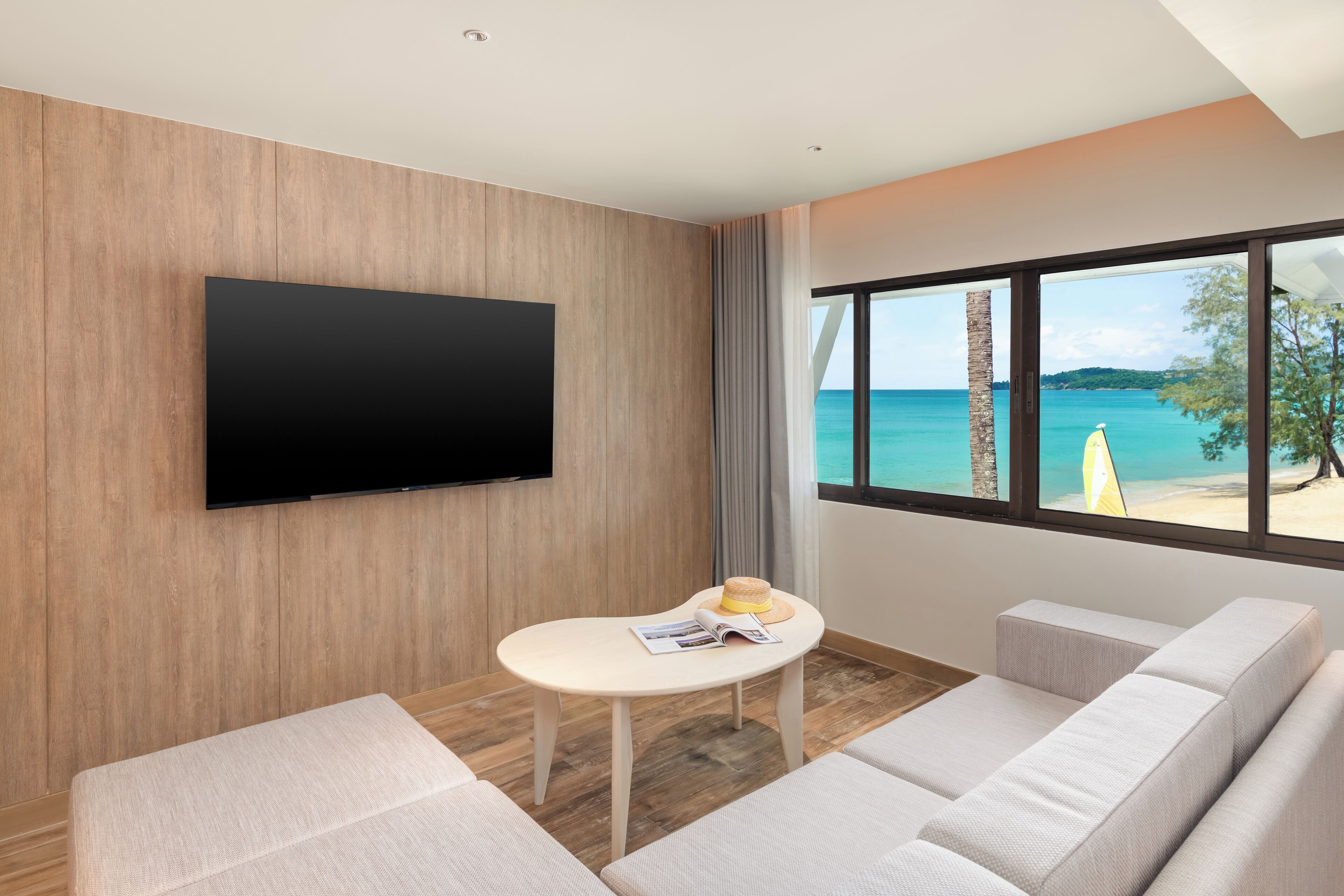 A modern hotel room with a large TV, a small table with a hat and magazine, and a view of the beach.