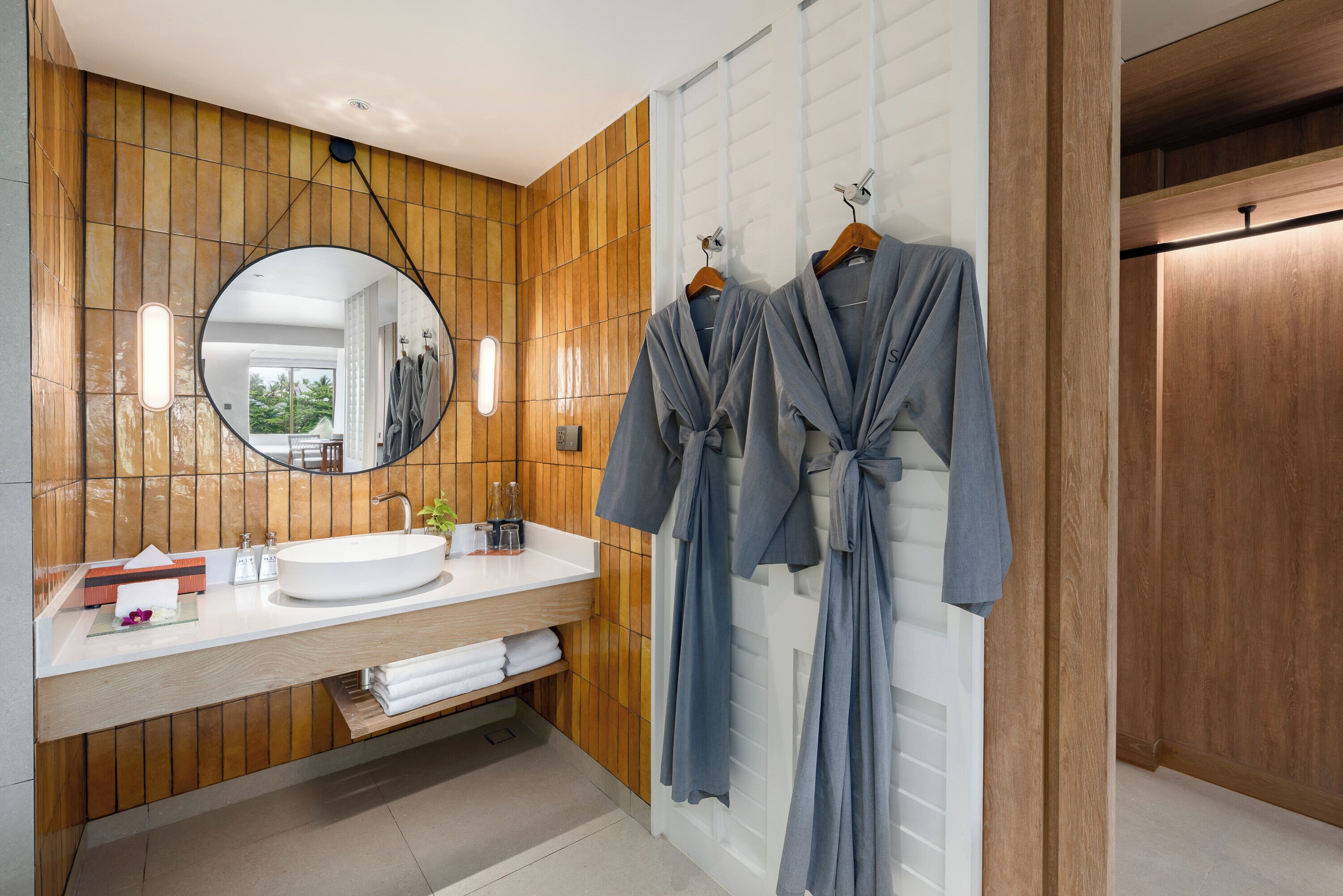 A modern bathroom with a wooden panel wall, a round mirror, a white sink, and two blue robes hanging on hooks.