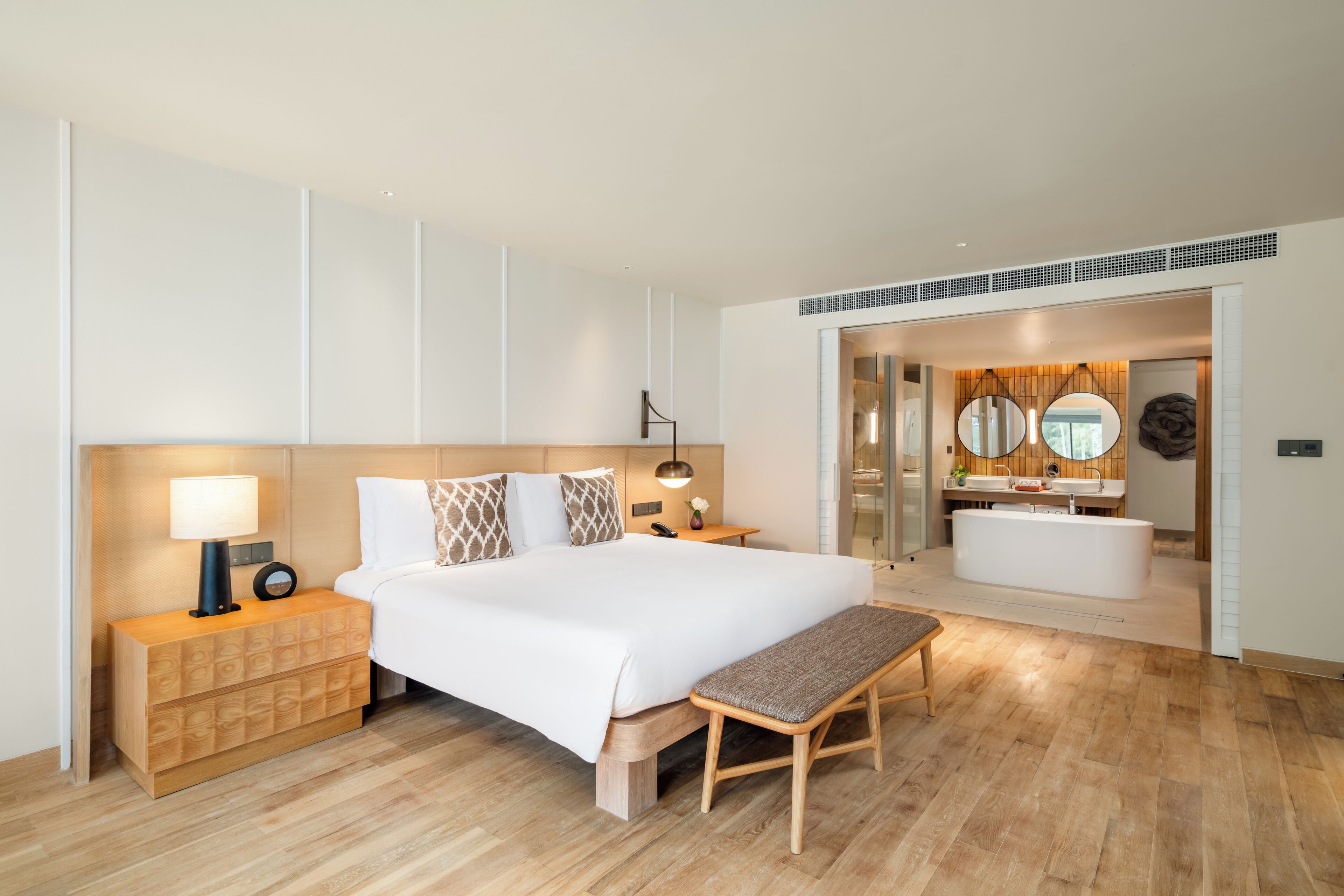 Una habitación de hotel moderna con una cama grande, mesitas de noche, un banco, un baño con bañera y un espejo.