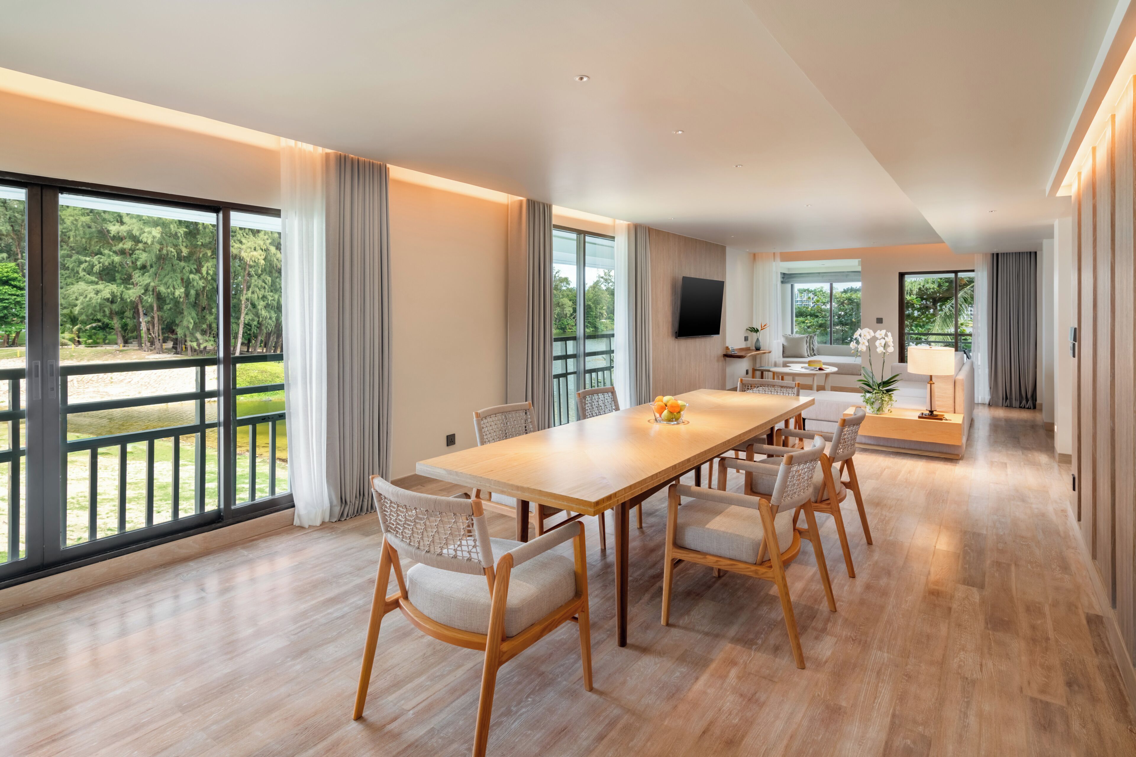 Un vaste espace dédié à la salle à manger, avec une longue table en bois et des chaises, de grandes fenêtres ornées de rideaux et une vue sur la verdure environnante.