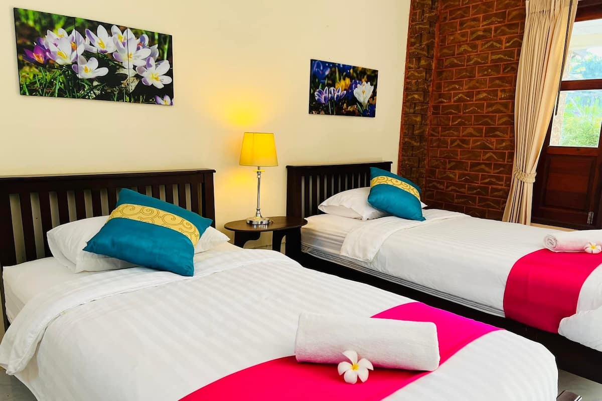 A hotel room with two beds, each with white bedding, blue and yellow pillows, and red and pink decorative pillows. There is a small table with a lamp between the beds, and a window with curtains.