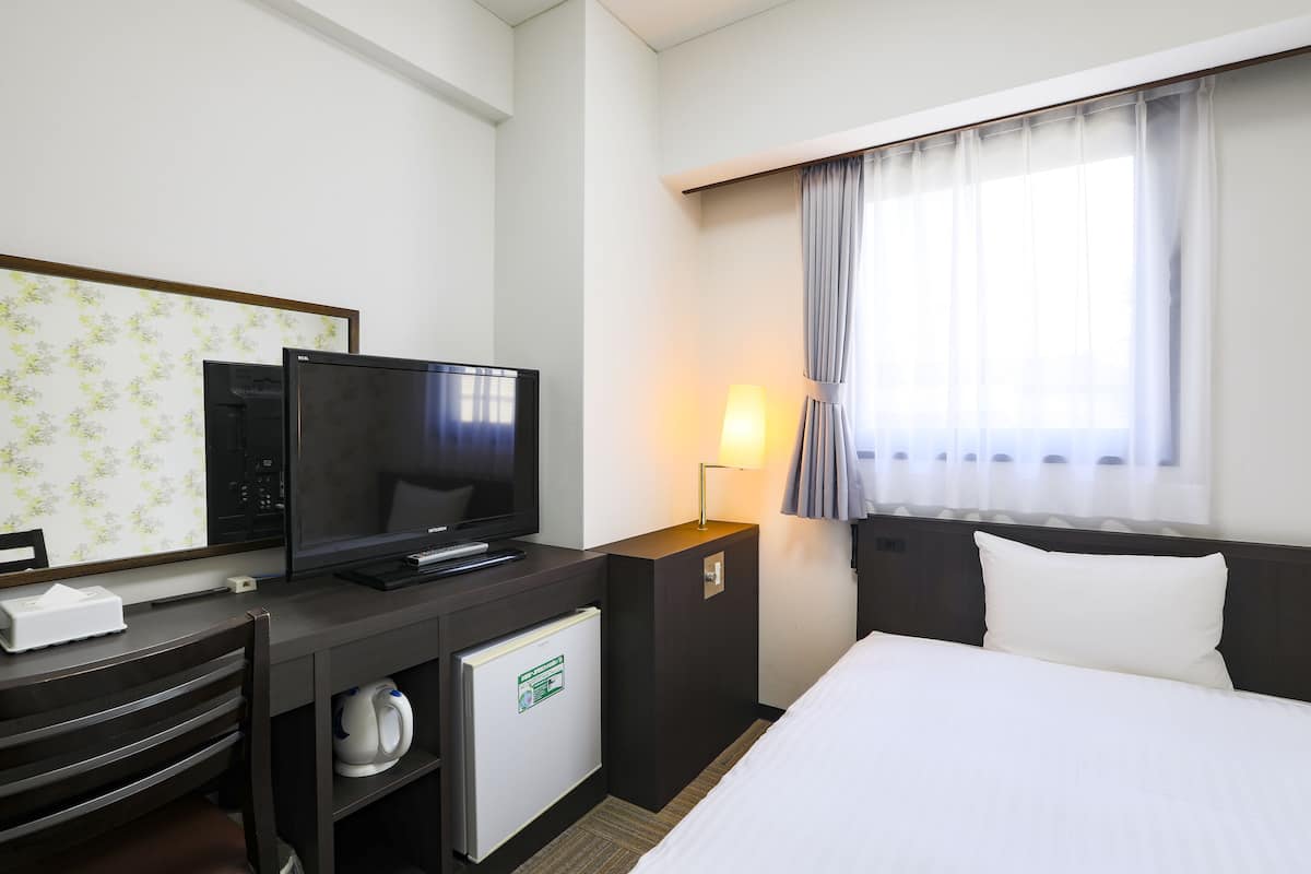 A hotel room with a bed, a TV, a mini-fridge, a desk with a lamp, and a window with curtains.