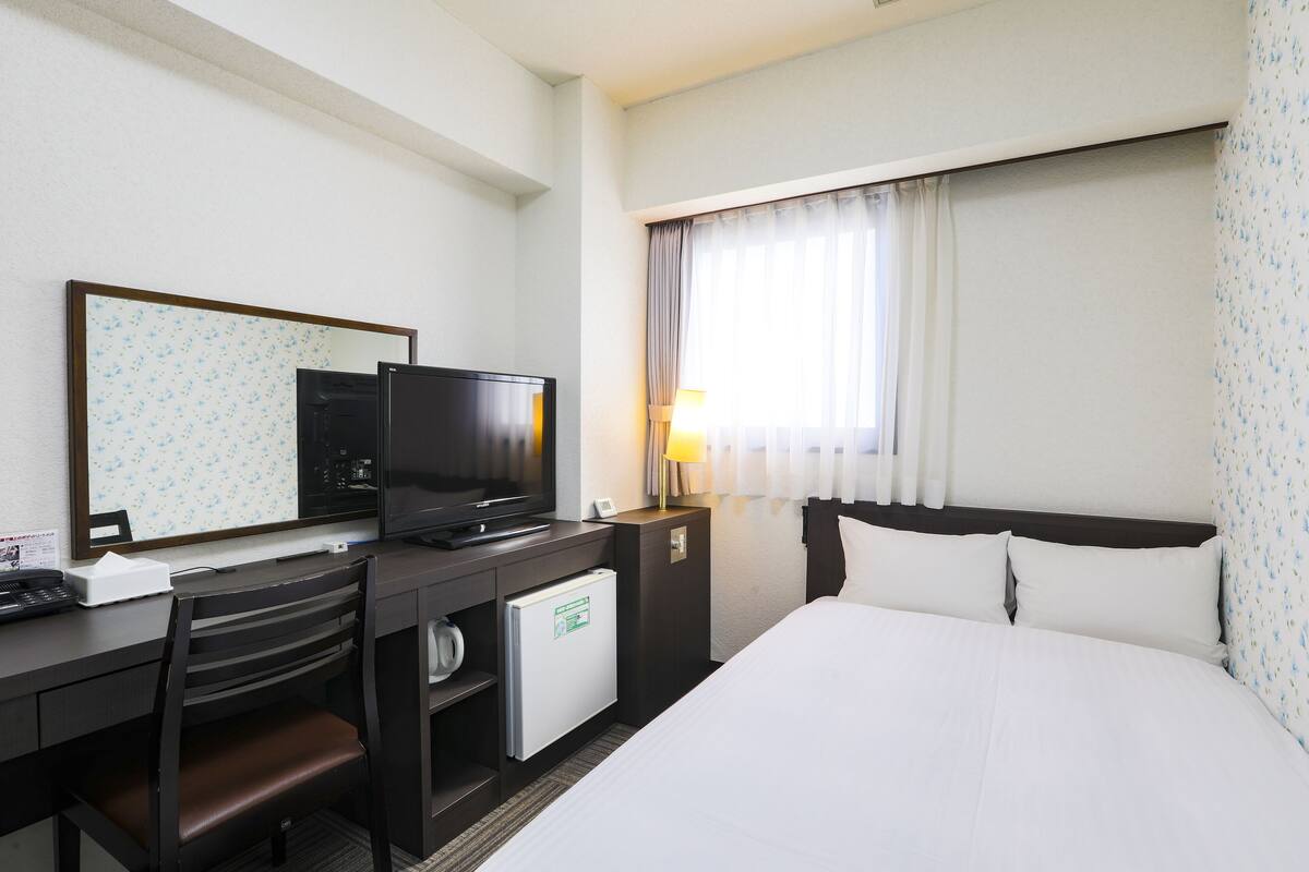 A hotel room with a bed, a desk with a television, a small refrigerator, a chair, and a window with curtains.
