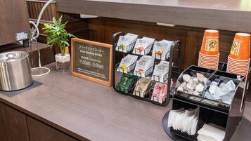 Zona de recepción de hotel con una máquina de café, sobres de té y café, y un cartel que indica bebidas ilimitadas disponibles de 11:00 a 0:00.