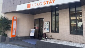 Un bâtiment sur lequel on peut lire l’inscription « KOKO STAY » et qui comprend un espace de détente avec un panneau indicateur.
