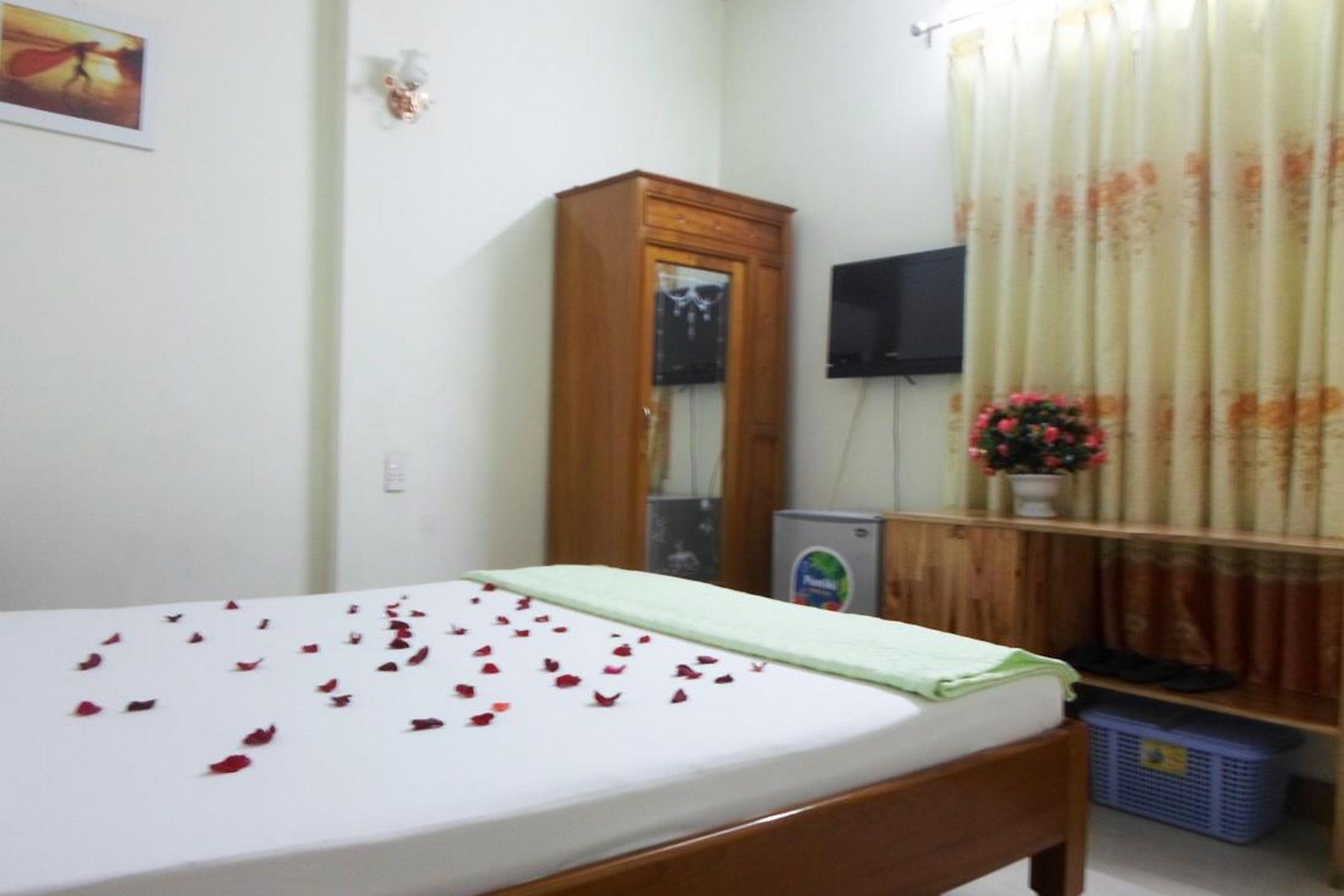 A bedroom with a bed, a wooden wardrobe, a television, a small refrigerator, and a window with curtains.