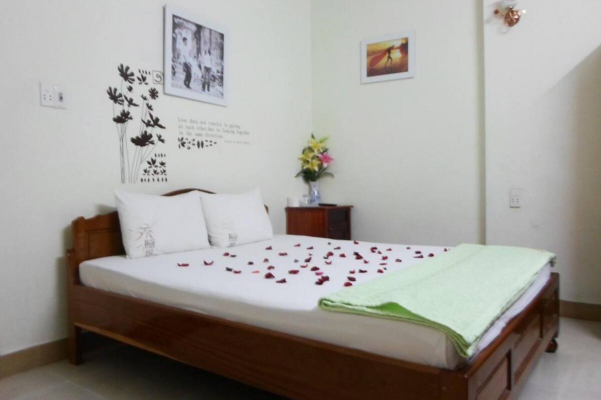 A neatly made bed with a green blanket, a wooden headboard, and white pillows. There is a small wooden nightstand with a vase of flowers and a framed picture on the wall above the bed.