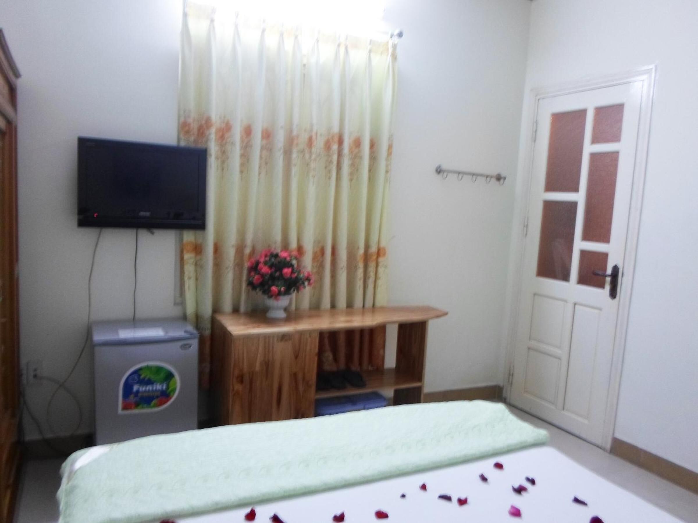 A room with a bed, a television mounted on the wall, a small wooden table with a vase of flowers, and a door.