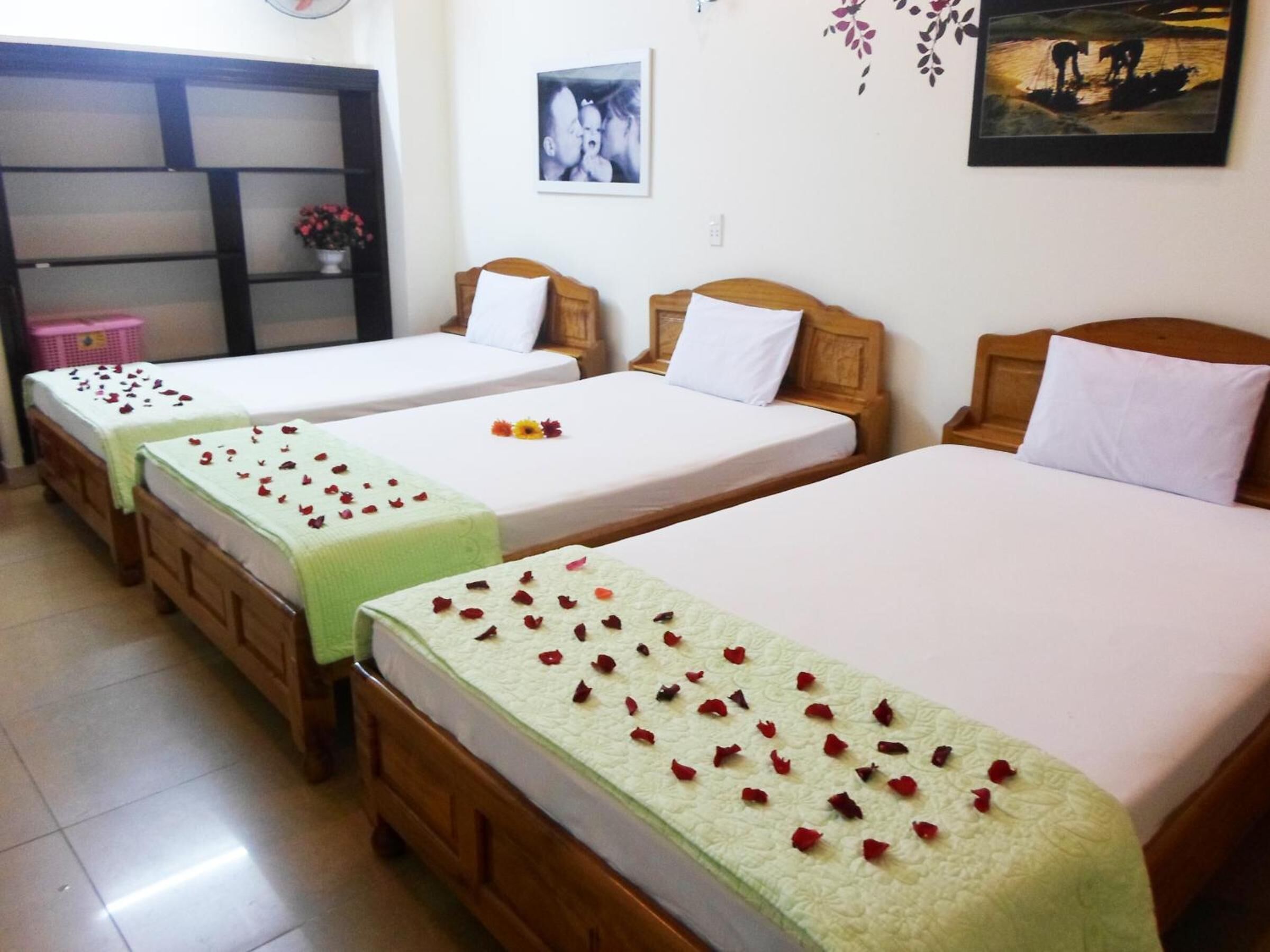 Une chambre d’hôtel avec trois lits simples, chacun recouvert d’une couverture verte et agrémenté de pétales de rose rouges. Le lit situé à droite est surmonté d’une tête de lit en bois, et à gauche, on trouve une petite armoire en bois avec un panier rose.