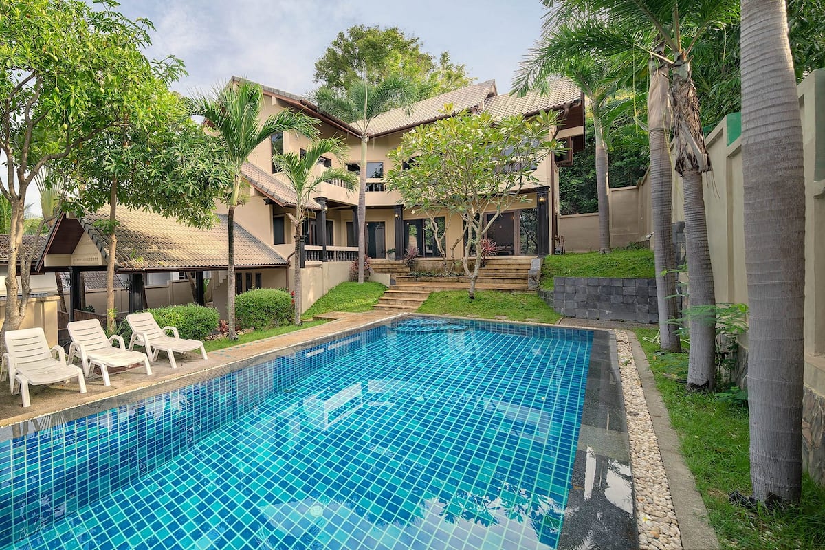 A swimming pool with lounge chairs, surrounded by palm trees and a multi-story building with balconies.