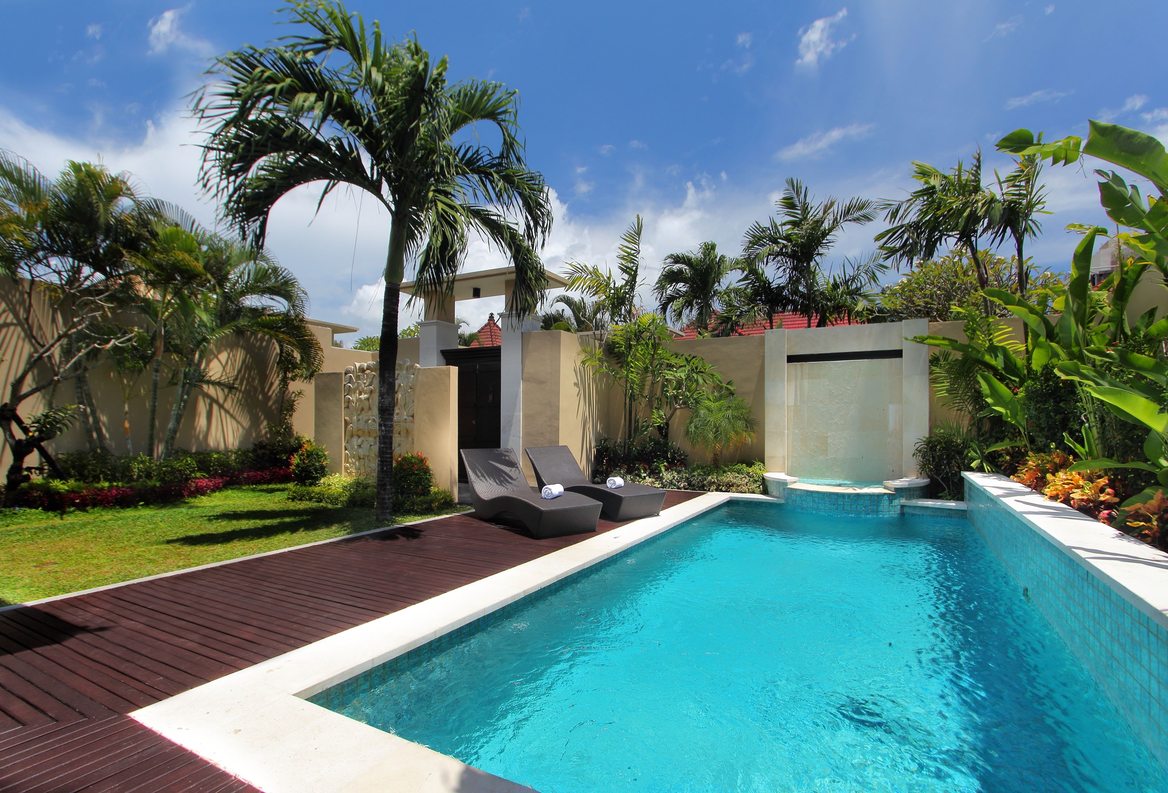 A swimming pool with two lounge chairs and a small hot tub, surrounded by palm trees and a wooden deck.