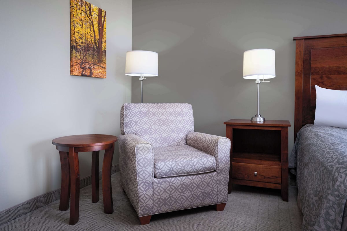 Habitación de hotel con un sillón estampado, dos mesitas de noche con lámparas, una cama y un cuadro en la pared.
