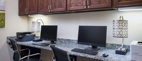 A workspace with two desks, each with a computer, keyboard, and mouse. There are two office chairs, a printer, and a lamp.
