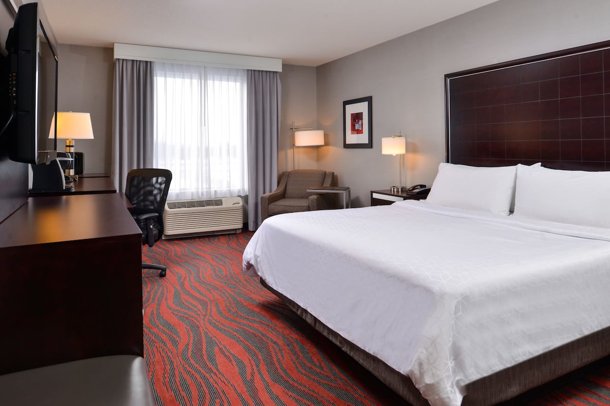 Ein Hotelzimmer mit einem großen Bett, einem Schreibtisch mit Fernseher, einem Sessel, einer Schreibtischlampe und einem Fenster mit Vorhängen.