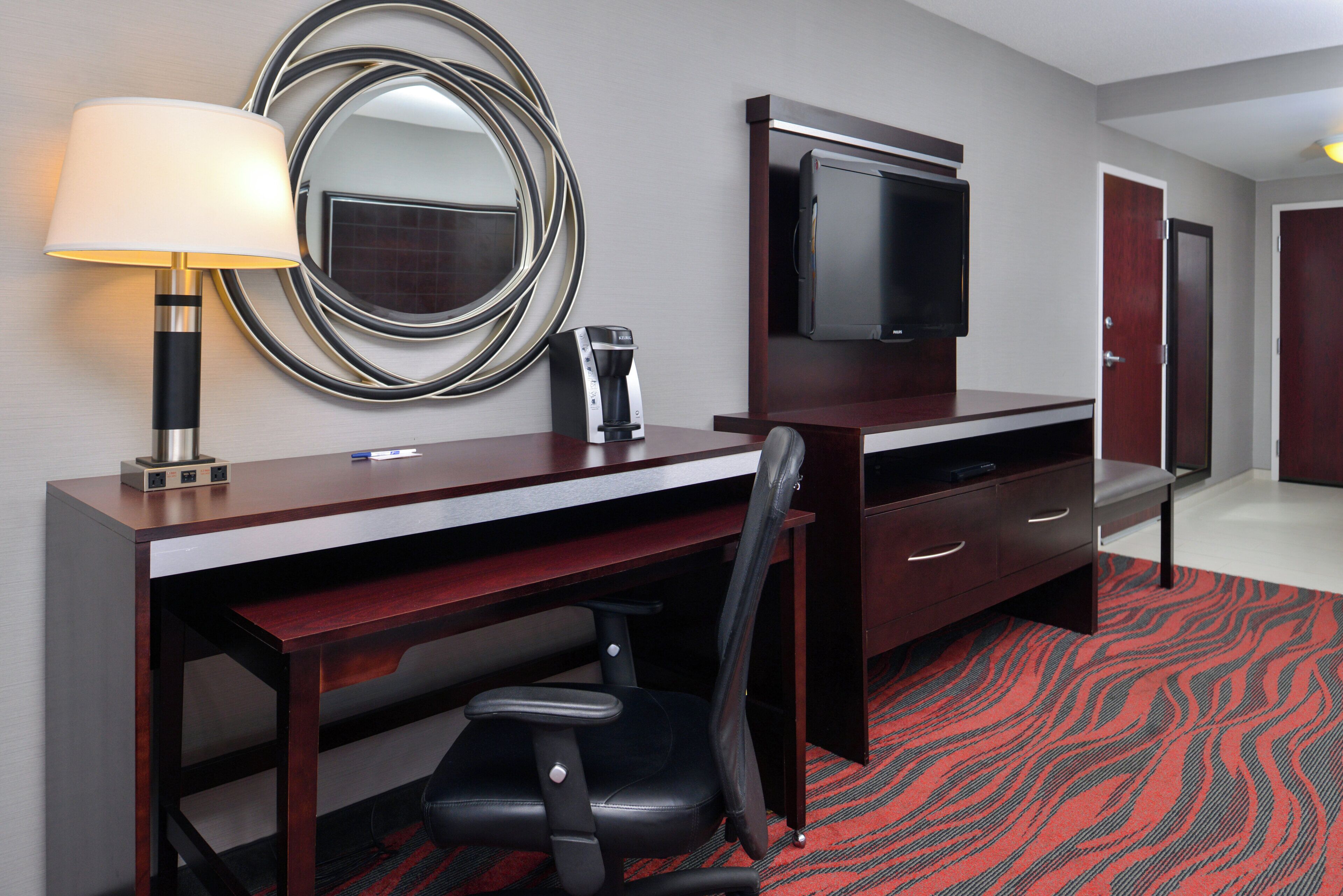 A hotel room with a desk, chair, lamp, coffee maker, TV, and a mirror.