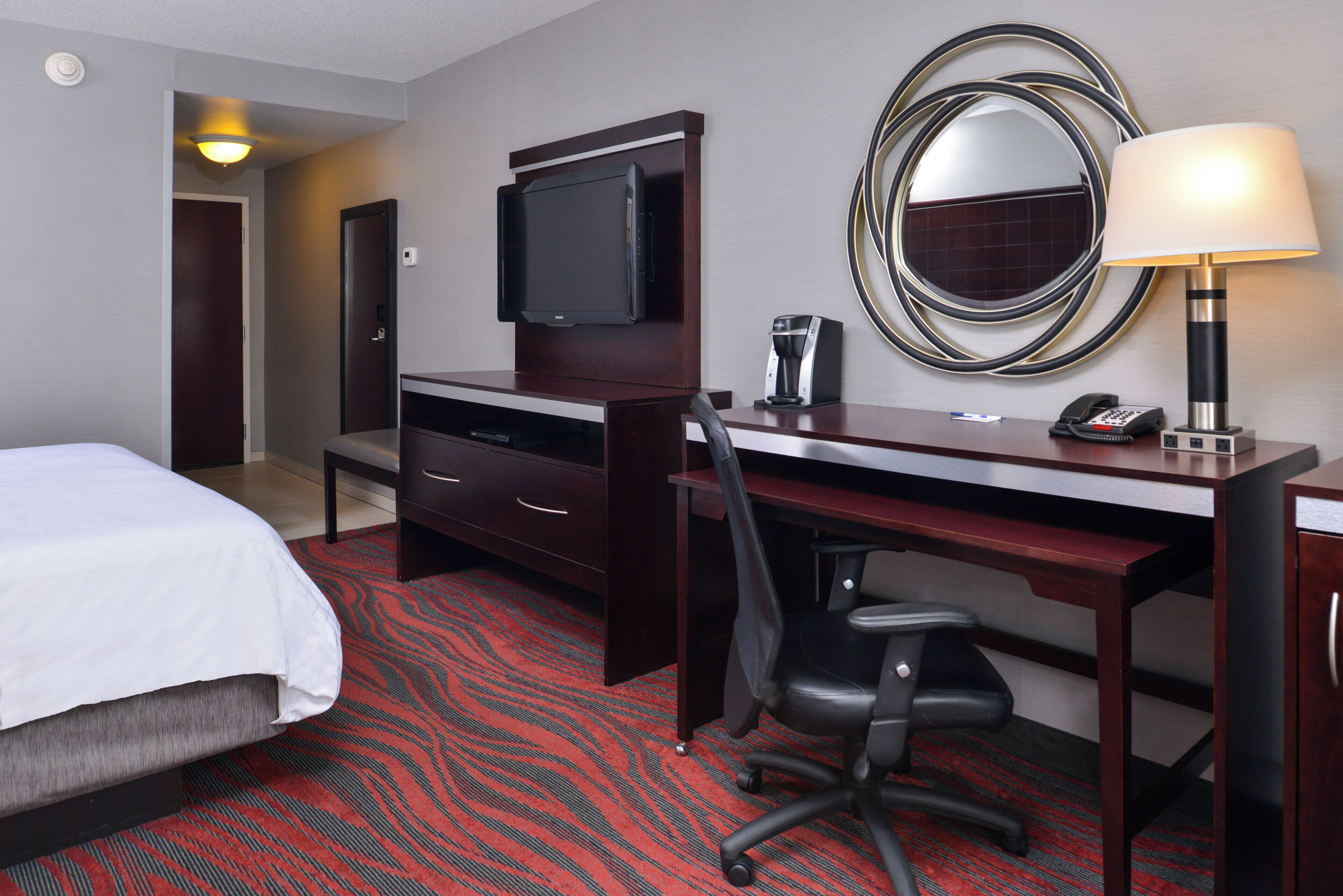 A hotel room with a bed, a desk with a chair, a television, a coffee maker, a telephone, and a mirror.