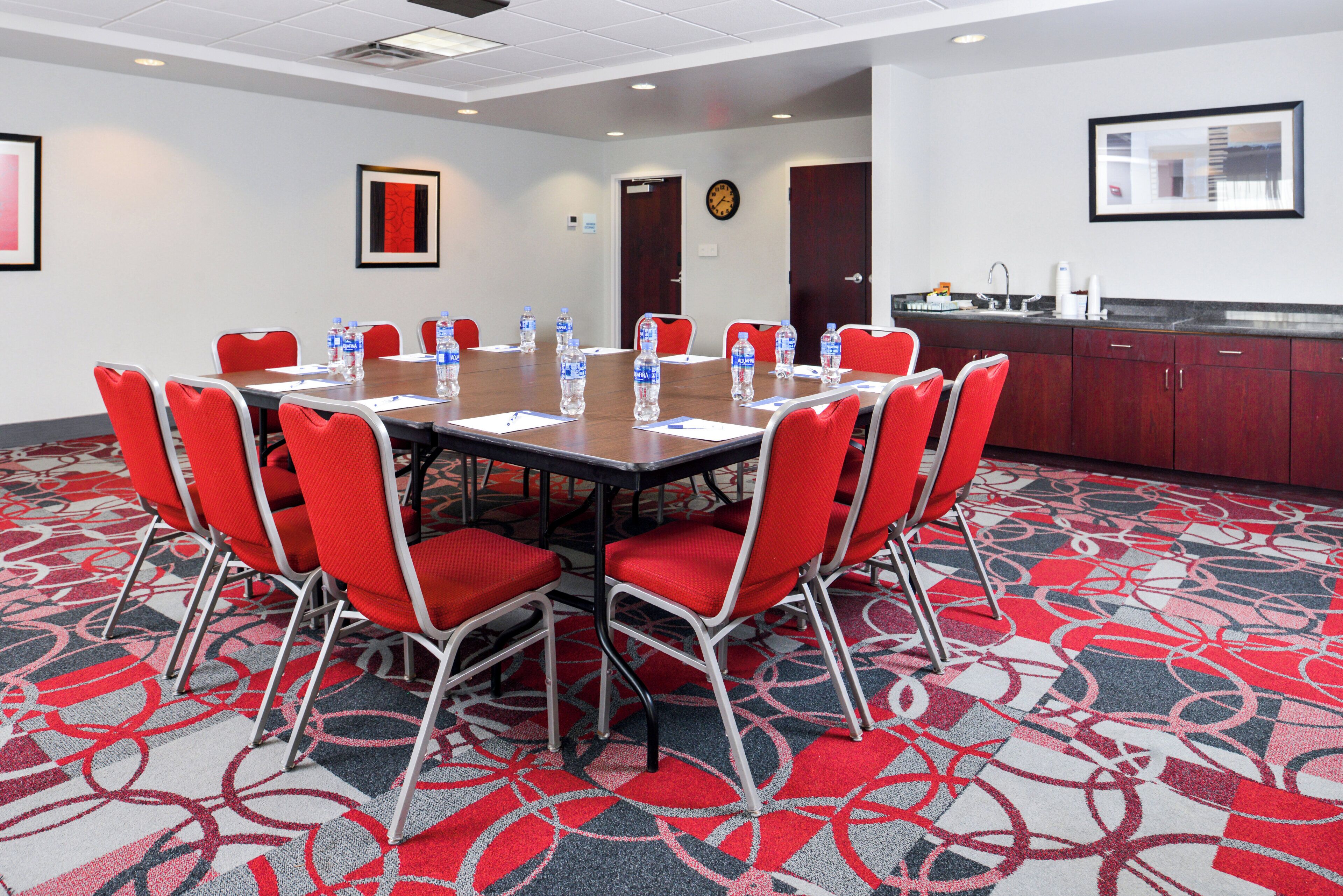 A conference room with a round table, red chairs, water bottles, and a clock on the wall.