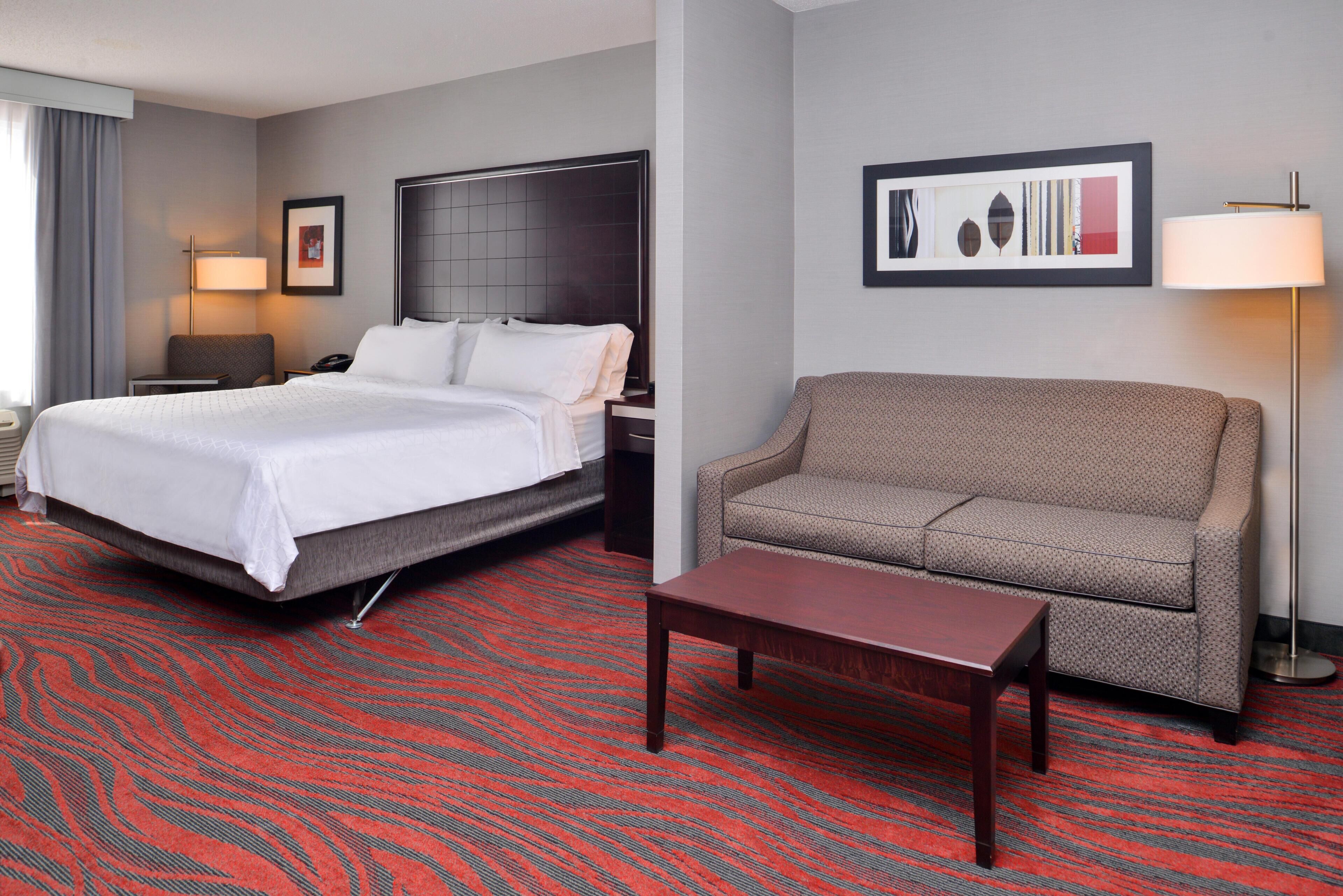 A hotel room with a large bed, a sofa, a small table, and a nightstand with a lamp.