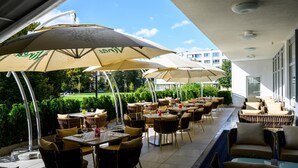 An outdoor dining area with tables and chairs, covered by umbrellas, and a view of a park and buildings in the background. - Lux Hotel (Banska Bystrica)
