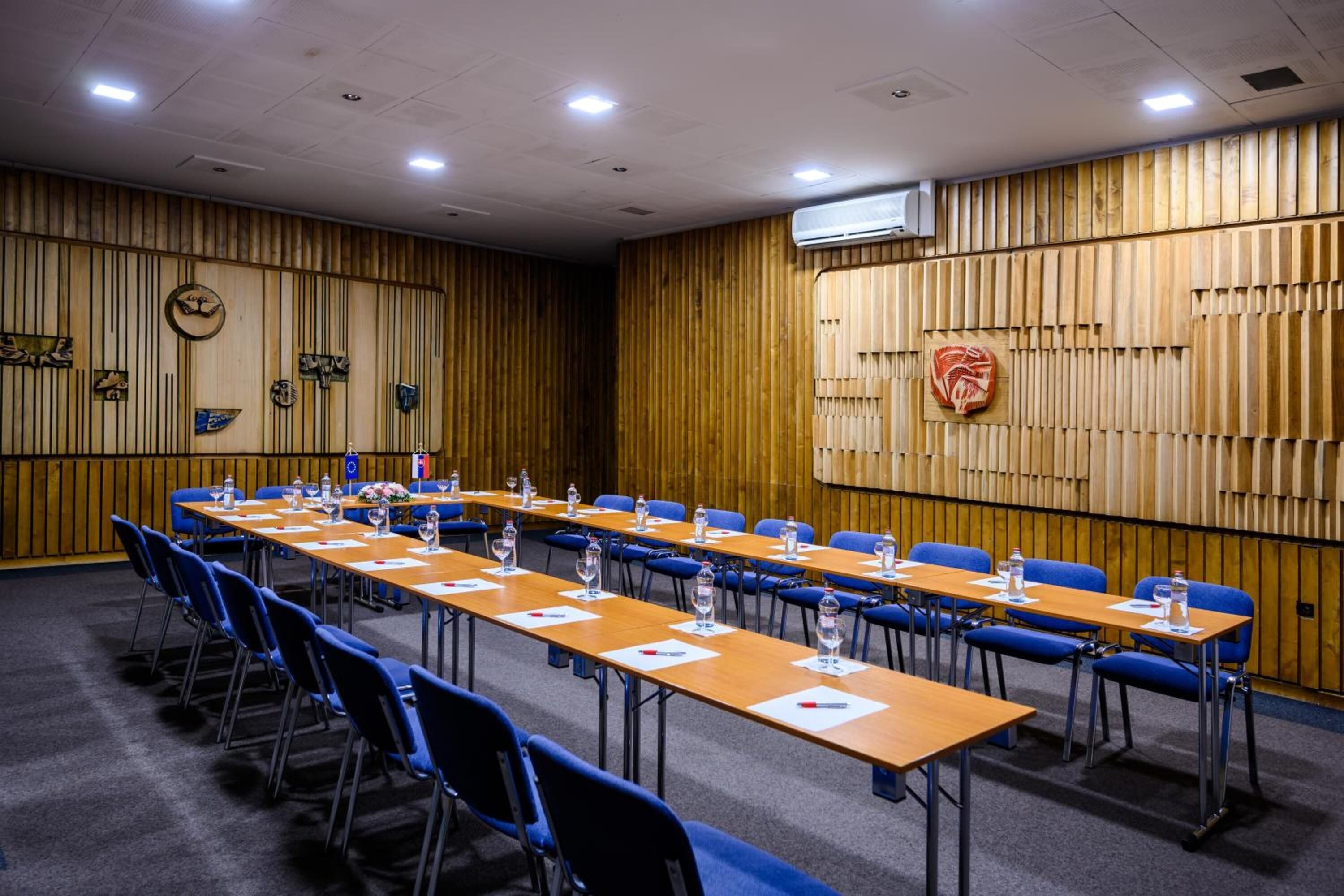Un salón de conferencias con mesas largas, sillas azules y un panel de madera decorativo.
